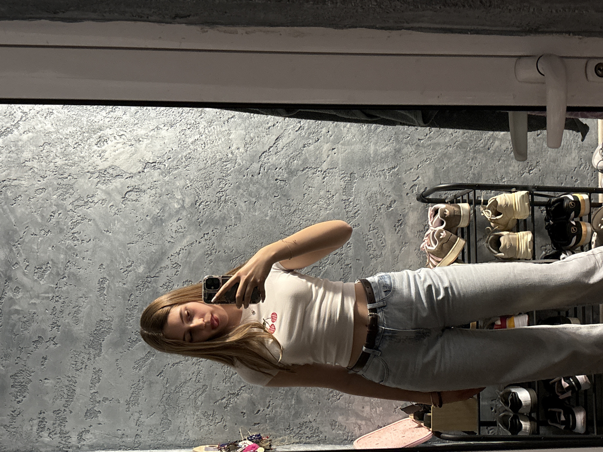 A young woman with long blonde hair is taking a selfie in a mirror. She is wearing a white cropped t-shirt with a cherry design, light-wash jeans, and a brown belt. The background shows a textured gray wall, a shoe rack with various pairs of sneakers, and a window frame with a white handle. The location cannot be determined solely from the visual information in the image; there is nothing that indicates the city of Panagyurishte, Bulgaria.