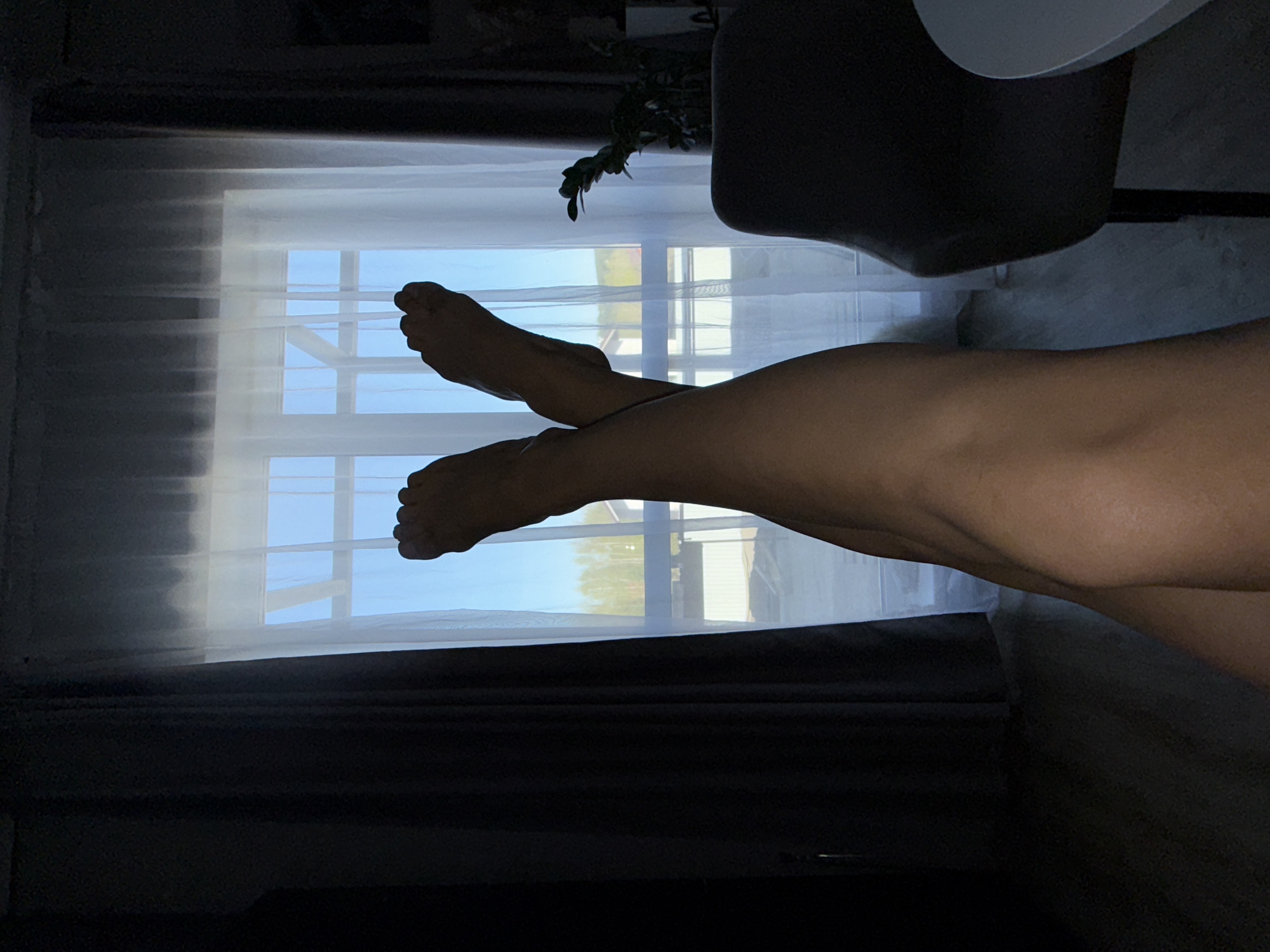 The image shows a pair of legs crossed in front of a window. Outside the window, there are trees and a building, possibly in Лобаново, Russia. A chair and a plant are also visible in the scene.