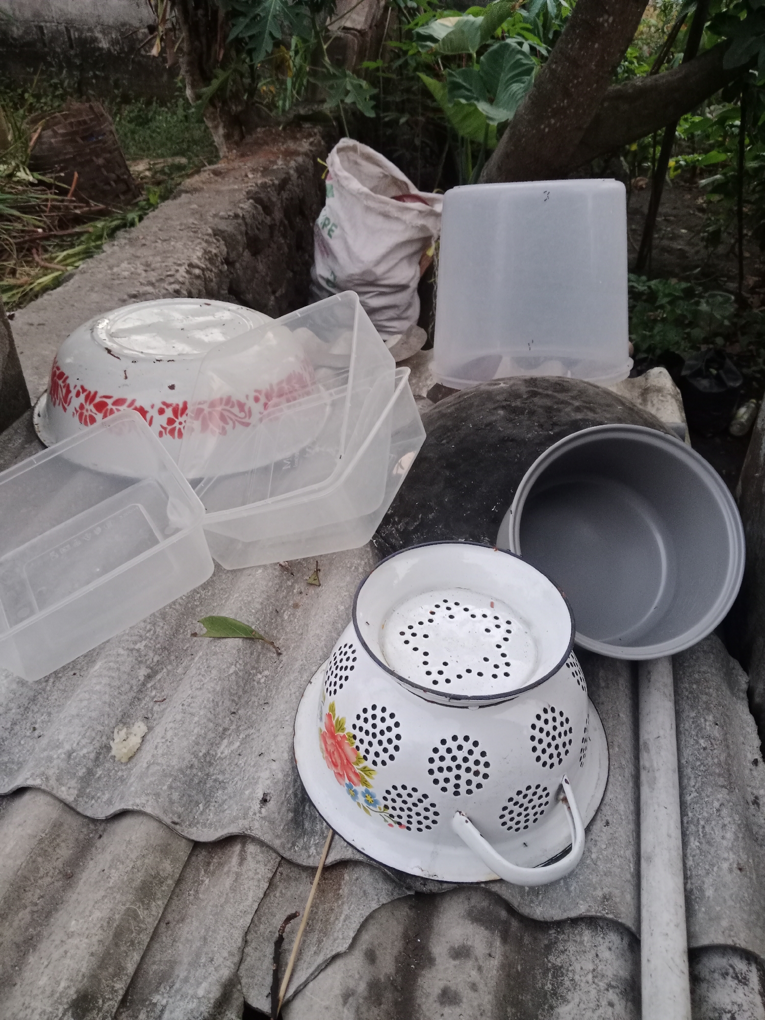 The image depicts an outdoor scene in Sumberasri, Indonesia, showing a collection of discarded household items and debris. In the foreground, a white enamel colander with a floral pattern and black trim sits on a corrugated surface, possibly roofing material. To its right is a gray pot, also likely discarded. Behind these items, several clear plastic containers and a white bowl with a red floral pattern are visible. A large white sack with some lettering is also present, leaning against a rough-surfaced object. The background consists of dense foliage and a tree trunk, suggesting a natural or overgrown area. The overall impression is one of disuse and neglect, with items left exposed to the elements. There are no people or animals in the frame.