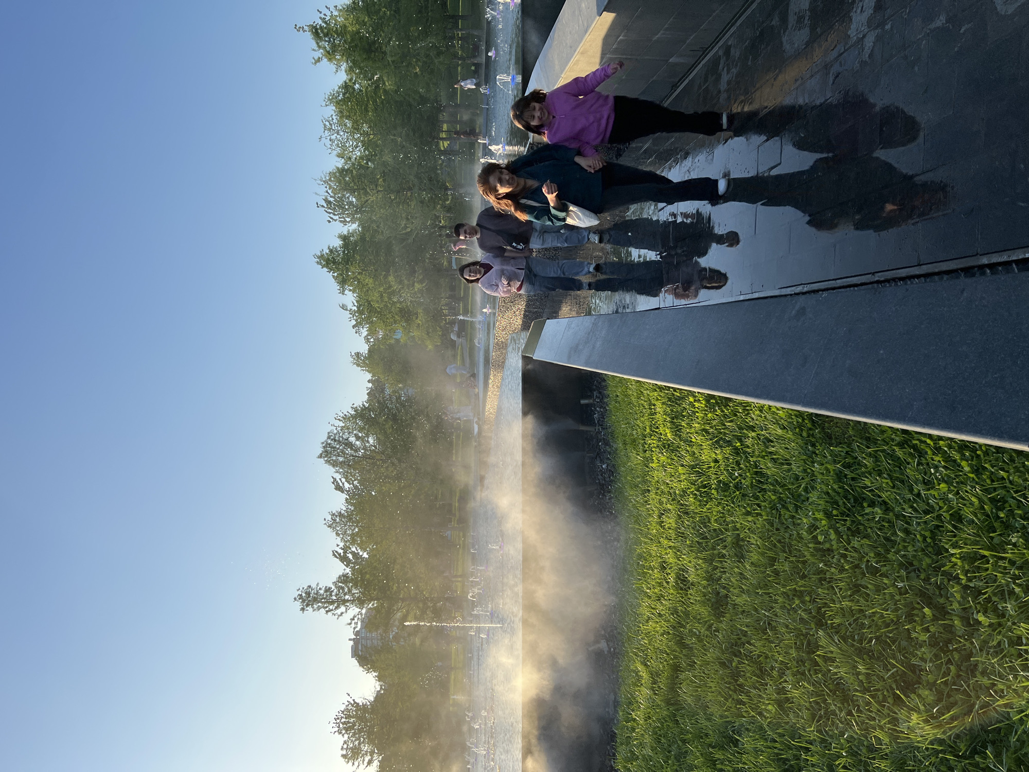 Four individuals stand on a wet, dark stone surface, their reflections visible. A woman in a purple jacket is on the right, while another in a teal jacket leans toward her. Two other people stand to their left. A fountain with a light mist and water jets is visible in the background, along with green grass and trees. The sky is clear blue. The location is likely the Krasnodar Park in Krasnodar, Russia, known for its fountains and recreational areas.