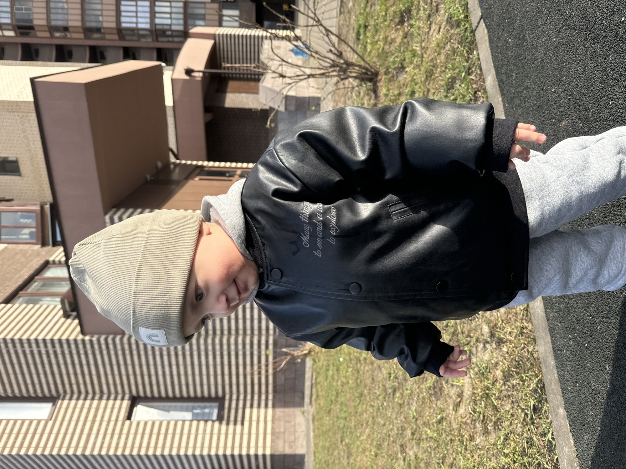 A young child is captured standing outdoors in Akulovo (Gp Kubinka), Russia, on a bright, sunny day. The child, likely a toddler, is wearing a light beige ribbed beanie, a black leather-look jacket over a grey hooded sweatshirt, and grey sweatpants. Their face is partially visible, with fair skin and dark eyes, looking downwards and slightly to the left, suggesting engagement with something on the ground.
The black jacket features white embroidered text on the left chest, which reads, "Many things to see and to explore."
The setting includes a patch of dry grass bordered by a dark grey or black paved surface, possibly a playground or walkway. Strong sunlight creates distinct shadows, indicating a clear, daytime environment, likely midday or early afternoon. In the background, several modern residential buildings are visible, displaying various architectural textures including striped and plain facades with numerous windows. Bare tree branches are also present, suggesting either early spring or late autumn. The overall impression is one of a child exploring their surroundings in a residential outdoor space.