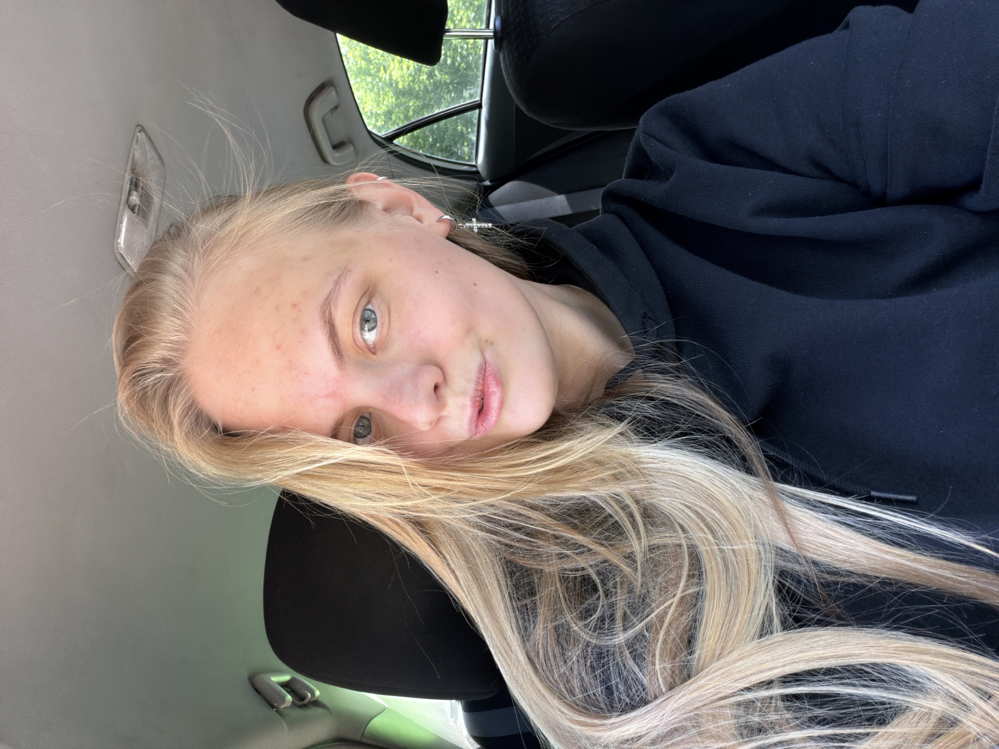 A young, light-skinned female with long blonde hair is seated inside a vehicle, possibly a car. She wears a black hoodie. Her face is angled towards the camera, and her eyes appear light-colored. A silver cross earring is visible on her left ear. The interior of the vehicle is visible, including the gray ceiling, a black seat headrest, and glimpses of the outside through the window, revealing green foliage. The image location is not explicitly identifiable as Chaikovsky, Russia, from the given visual information.