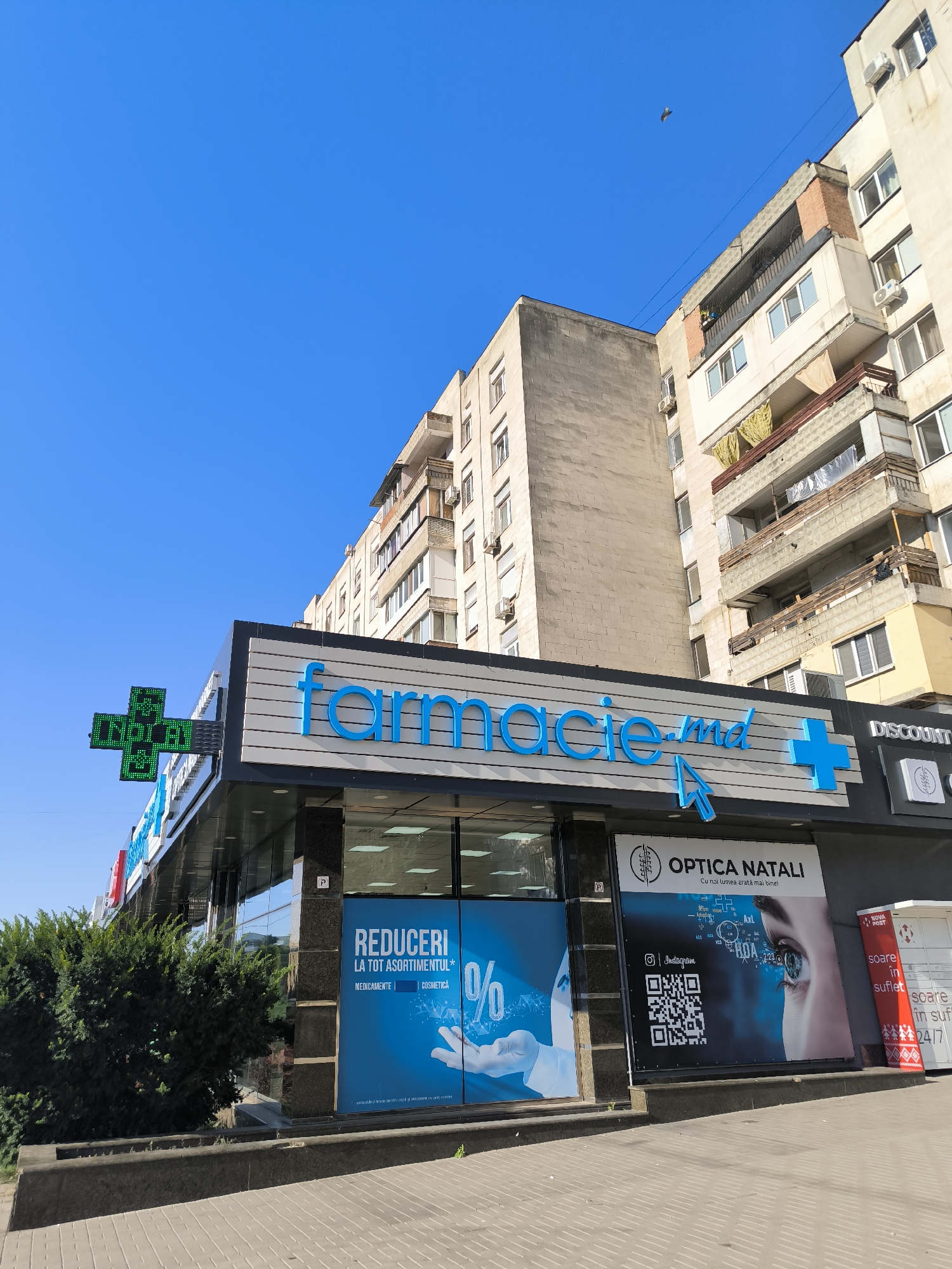 The image captures a street-level view of a commercial building in Chișinău, Moldova, under a clear blue sky. Dominating the storefront is a large sign for "farmacie.md," a pharmacy, with bright blue lettering against a striped gray background. To the left, a green illuminated cross sign, typical of pharmacies, is visible. Adjacent to the pharmacy's entrance is a store labeled "OPTICA NATALI," featuring a large advertisement with an image of a human eye and text in Romanian. A Nova Post parcel locker is also present on the right side of the building. The architecture suggests a Soviet-era residential building in the background, with multiple balconies and windows. The foreground shows a paved sidewalk and a small, dense green hedge. The overall scene is one of a commercial street in a city, likely during daytime.