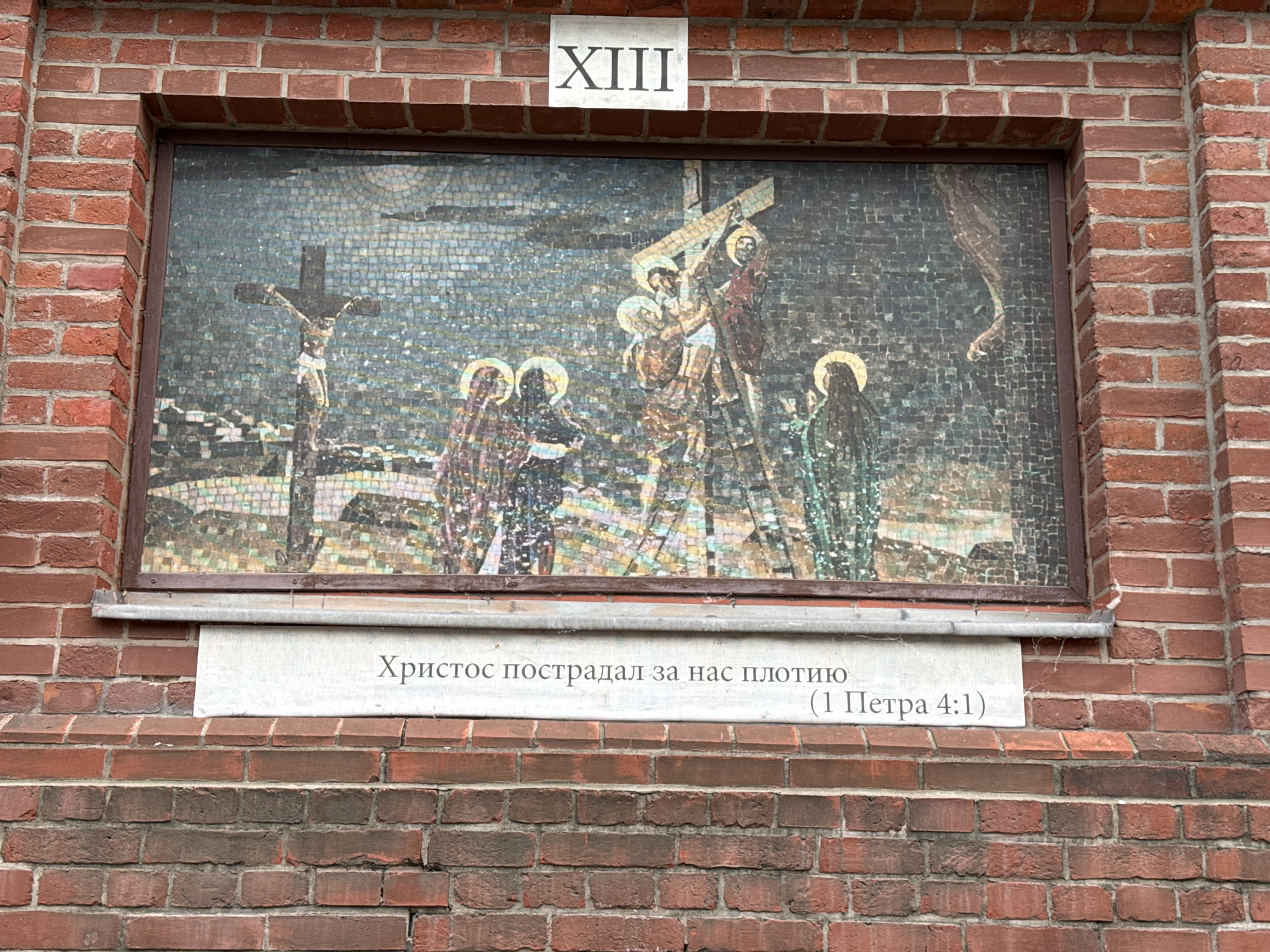 This is a close-up, eye-level, full outdoor shot of a brick wall with a mosaic artwork above a stone plaque. The mosaic depicts a scene from the crucifixion of Jesus Christ, with several figures surrounding him on crosses. The artwork is framed by a dark border and set into the brick wall. Above the mosaic, a rectangular sign with Roman numerals "XIII" is affixed to the brick. Below the mosaic is a light-colored stone plaque with text in Cyrillic, which appears to be a quote from scripture. The brickwork is a deep red-brown color. The overall lighting suggests it is daytime. The mosaic and plaque are mounted on a section of the brick wall that is slightly recessed compared to the surrounding bricks. The image does not provide specific geographical details within Moscow, Russia, but the style of the mosaic and inscription suggest a religious or historical context.