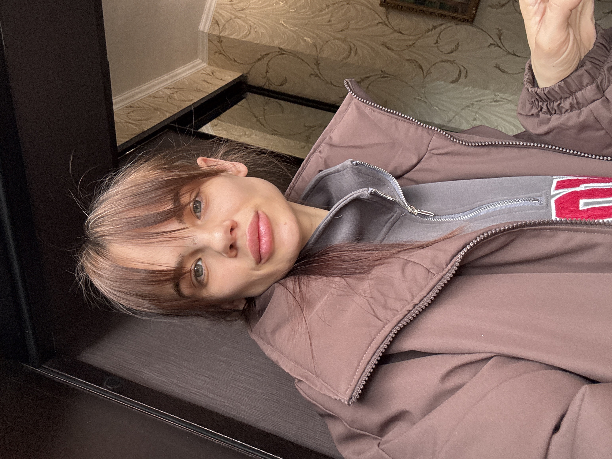 A woman is lying on a dark wooden surface, wearing a brown puffer jacket and a gray sweatshirt with a red design. She has brown hair with blonde highlights and is looking at the camera. The background includes decorative wallpaper and a dark, possibly glass, surface. The location is likely an interior space. The image does not definitively establish the location as Rostov-on-Don, Russia.