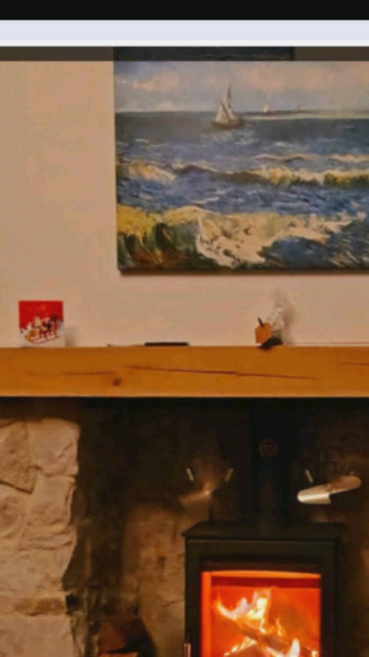 The image depicts a cozy indoor scene, likely a living room, featuring a fireplace with a wood-burning stove. Above the fireplace is a wooden mantelpiece adorned with a small red and white box, possibly a gift or decoration. A painting of a seascape with sailboats hangs on the wall above the mantelpiece. To the right of the mantelpiece, a small object, possibly a piece of packaging, rests. The fireplace itself has a stone facade, and the wood-burning stove has a glass door revealing a roaring fire. Two small metal fans are attached to the top of the stove, designed to circulate heat. The overall impression is one of warmth and comfort. The provided context does not include any information about Hongkou District, China, so this detail cannot be incorporated into the description.