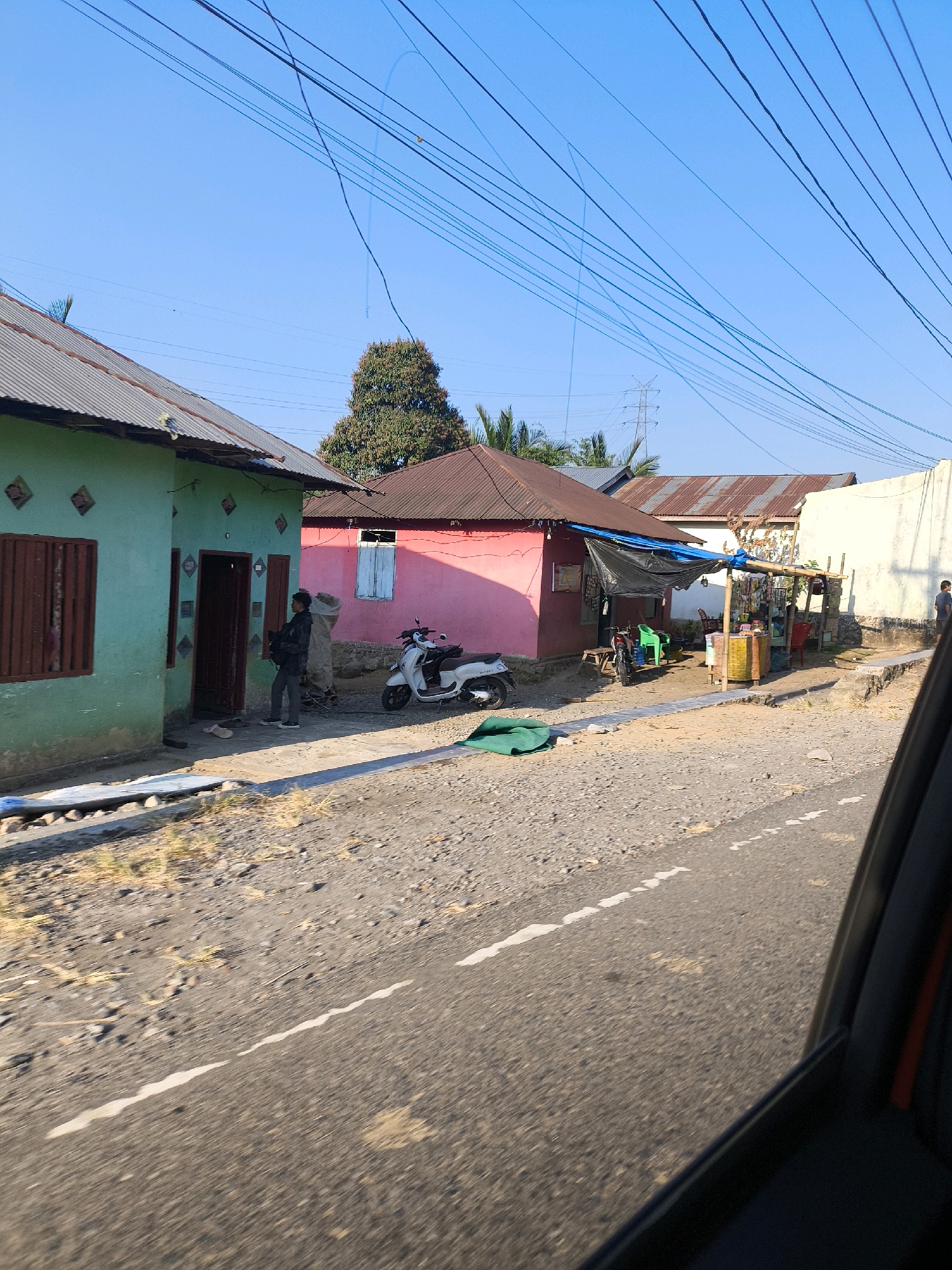 The image depicts a street scene in Dulupi, Indonesia, under a clear blue sky. On the left, a pale green house with a corrugated metal roof has a dark brown wooden door and a window with dark brown shutters. Two individuals are standing in the doorway of this house, one carrying a large beige bag. To the right of the green house, a pink building with a red-brown corrugated metal roof features a light blue shuttered window. A white scooter is parked in front of the pink building. Adjacent to the pink building, a small roadside stall is set up with various goods displayed on shelves and a yellow counter. A green plastic chair is visible near the stall, and a green tarp is lying on the ground in front of the pink building. The road in the foreground is paved with asphalt and has a white painted line. The surrounding area is characterized by gravel and sparse vegetation. Power lines are strung across the upper portion of the image.