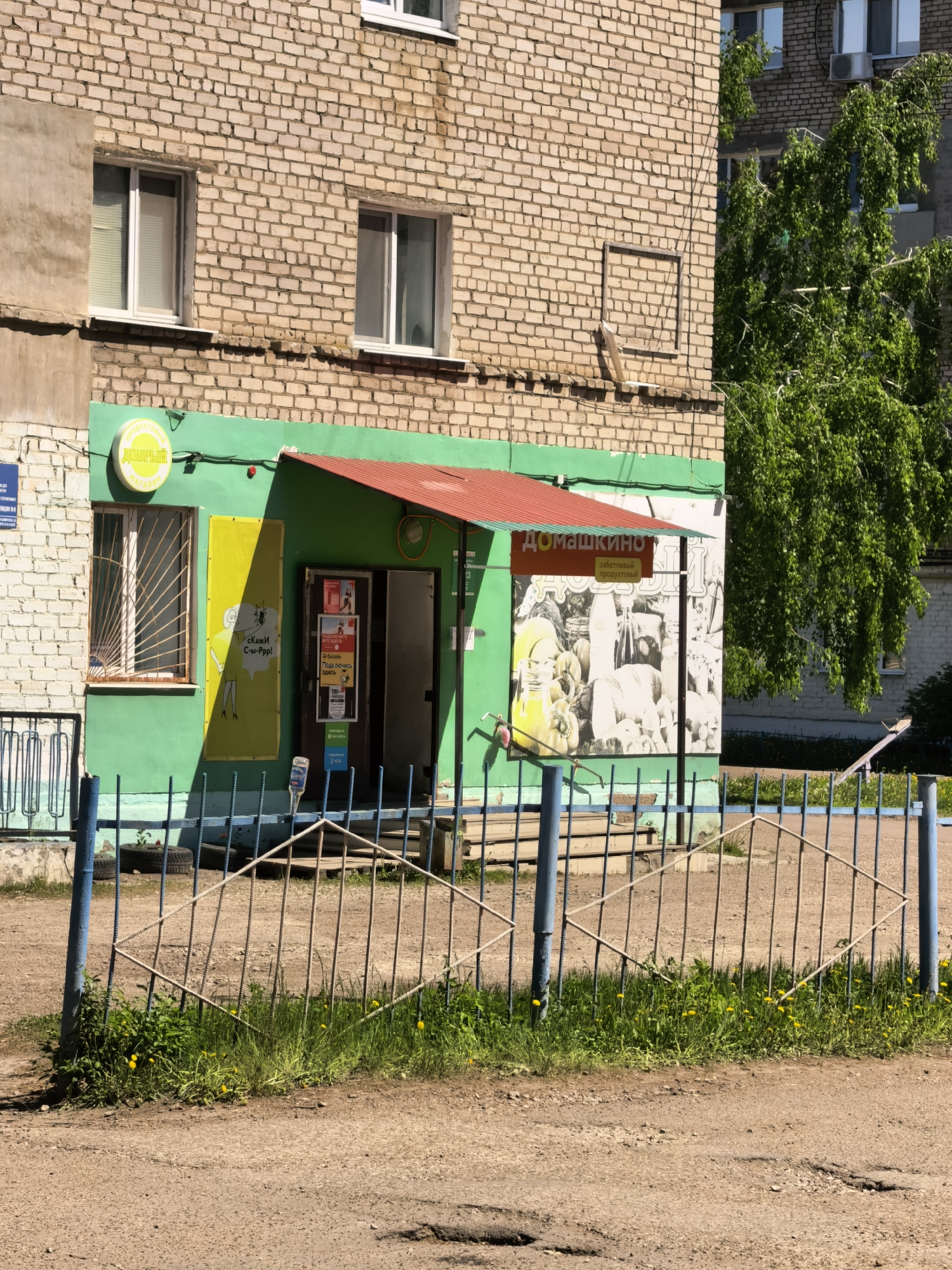 The image shows a ground-level view of a small retail store in Sterlitamak, Russia. The building is primarily constructed of light-brown brick. A portion of the building is painted green and contains signage for the store "Домашкино" (Domashkino), a заботливый продуктовый (caring grocery store). There is also signage featuring the word "ДОБРЫЙ" (Dobryy), which means "kind" in Russian, as well as marketing material showing a woman with a camera and the phrase "скажи C-ы-Ррр!" (Say Cheese!). A sign advertising 2 Билайн (Beeline) states, "Подключись здесь" (Connect here).
The entrance to the store is visible, and the area in front of the store is fenced off with a metal railing painted blue. The ground is a mix of dirt and grass, and there are dandelions growing near the fence.