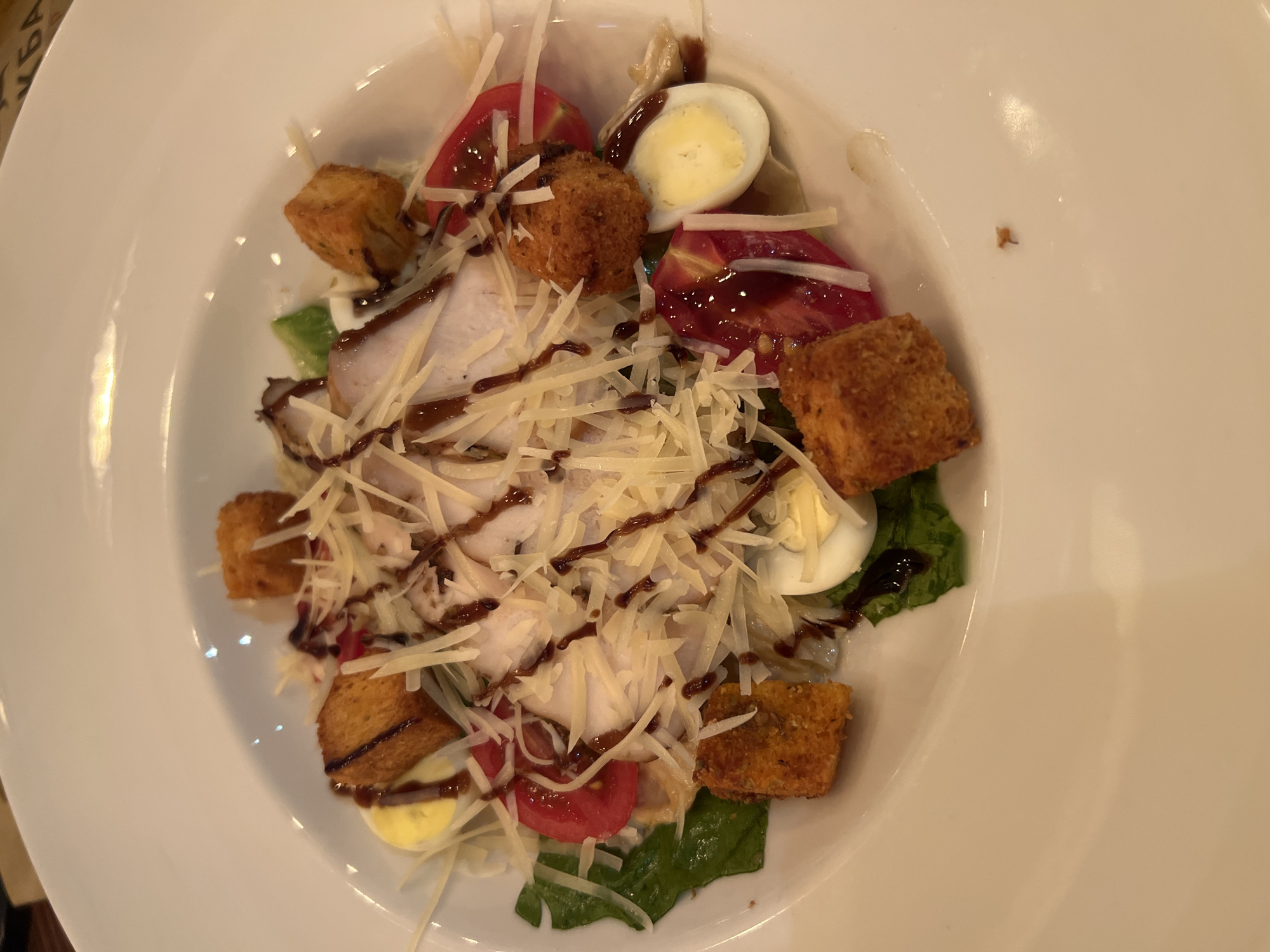 A close-up, overhead shot captures a white bowl of Caesar salad, presented on a white plate. The salad is comprised of lettuce, sliced chicken breast, cherry tomato halves, hard-boiled quail egg halves, croutons, and shredded Parmesan cheese. A dark, glossy dressing is drizzled over the top. The background reveals the edge of a white table and a portion of a menu with Russian text, hinting at the location as Novosibirsk, Russia. The lighting is dim and warm, suggesting an indoor dining environment.