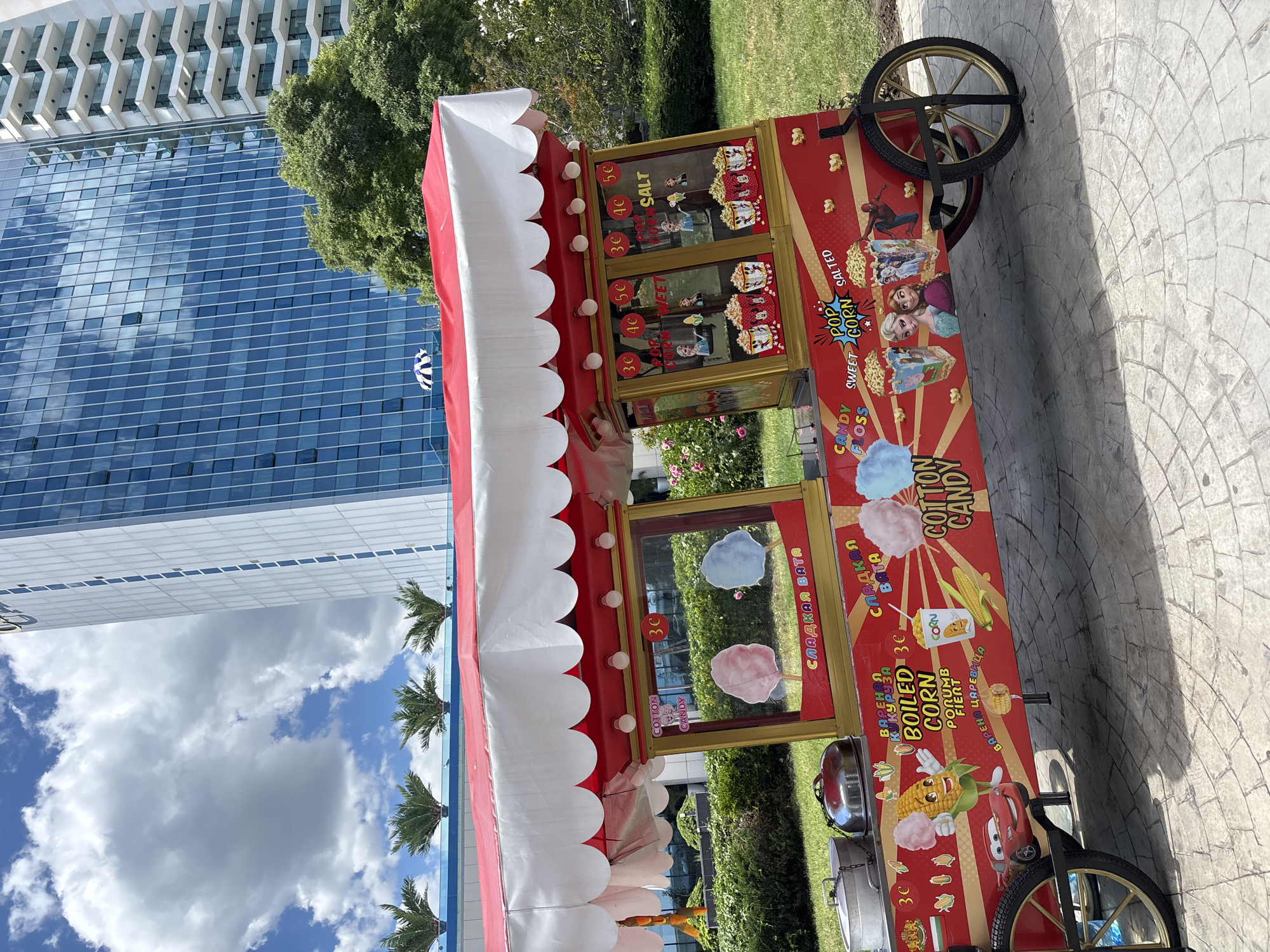 This image depicts a vibrant red and white food cart selling cotton candy and popcorn, situated on a paved area in front of lush green landscaping. In the background, a modern, tall glass-fronted building rises against a bright blue sky with scattered white clouds. The food cart is adorned with colorful cartoon characters, including a smiling corn cob, Lightning McQueen, and characters from Disney's Frozen, as well as images of popcorn and cotton candy. Text on the cart indicates prices in Euros and mentions "COTTON CANDY," "SWEET CORN," and "SALTED POPCORN," with some text in Cyrillic script. Two large silver pots are visible on the left side of the cart. The overall scene suggests an outdoor public space, likely a park or promenade, in Varna, Bulgaria, given the architectural style of the building and the presence of palm trees visible on a balcony in the background, which are commonly found in Varna's seaside resorts.