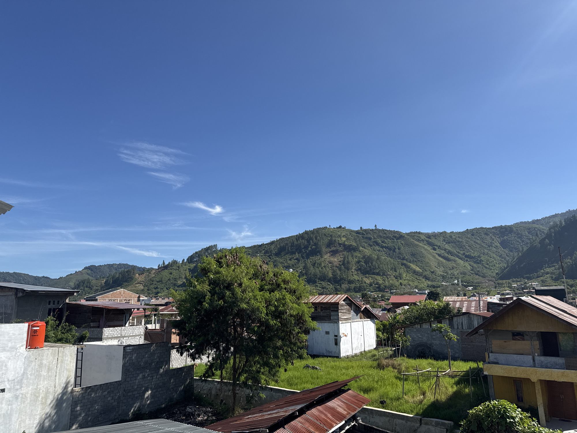 The image captures a view of the city of Kalalengkio, Indonesia, under a clear blue sky. The foreground features a variety of structures, including concrete walls, houses with corrugated metal roofs, and a prominent orange water tank. A large green tree stands to the left of center. In the midground, more buildings are visible, constructed from brick and wood, some with red roofs. The background is dominated by lush green, rolling hills and mountains that form a backdrop to the settlement. The scene appears to be a residential area situated in a valley. There are no people visible in the image.
