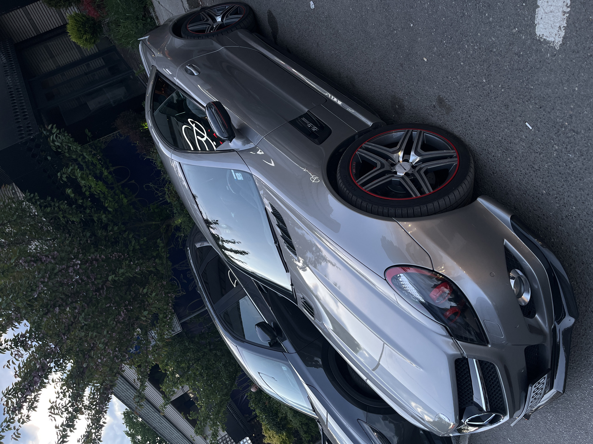 The image shows a gray Mercedes-Benz SLR McLaren parked on an asphalt street. The car has distinctive wheels with a red accent ring. It's positioned on the side of a road, with a building and foliage visible in the background. The location is Sofia, Bulgaria. Another car, which appears to be black, is parked directly behind the SLR. The sky is visible reflecting on the windshield. The overall environment suggests an urban street scene during daylight.