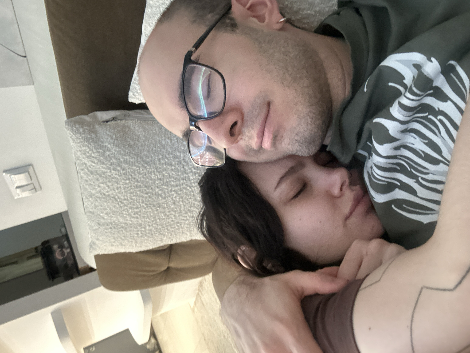 A couple is lying down indoors. A man with glasses, a bald head, and a beard is on top of a woman with dark hair. They are both sleeping. The man is wearing a green hooded sweatshirt with a white design. The woman is wearing a brown shirt and has tattoos on her arm. They are on a couch with patterned pillows. The location is presumed to be a residence in Khimki, Russia.