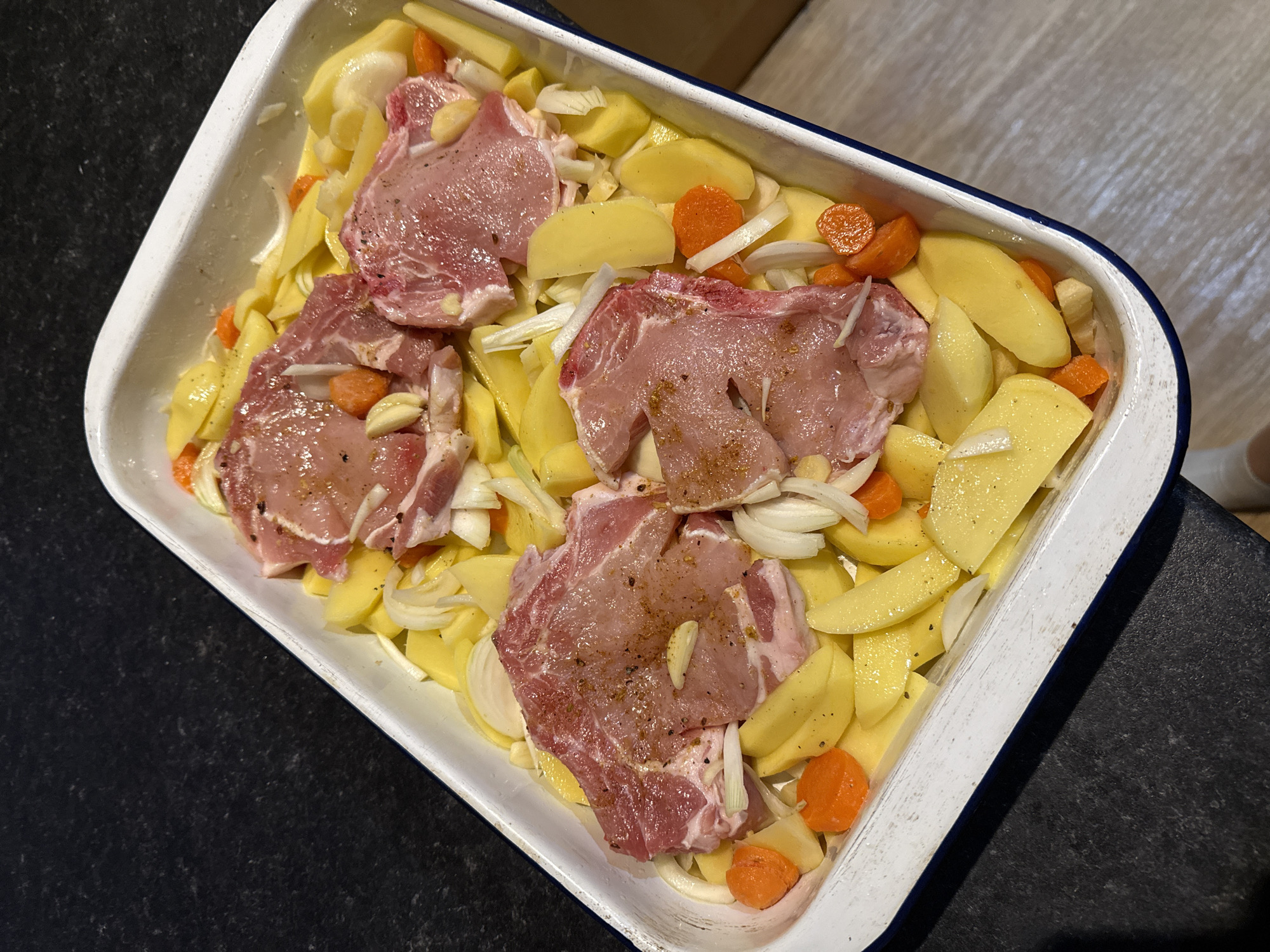 The image shows a white and blue enamel roasting pan filled with raw pork chops, sliced potatoes, carrots, and onions. Garlic cloves are scattered on top. The pan sits on a dark countertop. The location, Great Yarmouth, United Kingdom, is not directly indicated in the visual information of the image.