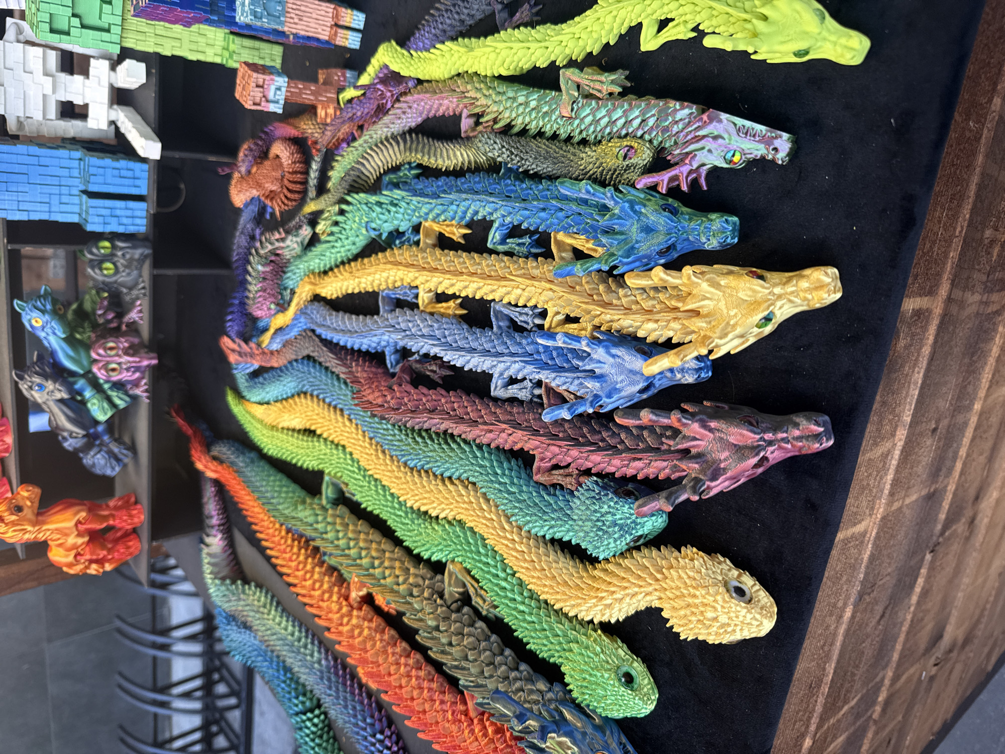 The image depicts a collection of colorful, segmented dragon toys displayed on a dark, textured surface, possibly a tablecloth or felt. The toys are arranged in rows and appear to be made of plastic, with a metallic sheen in various iridescent colors, including green, blue, yellow, red, and purple. Each dragon is articulated, with a detailed head, body, and tail, featuring individual scales and legs. In the background, other decorative items are visible, including what appear to be pixelated figures and a small orange horse-like figurine. The overall setting suggests a market stall or display for crafts or collectibles. The location is not identifiable from the image itself, therefore I cannot confirm it is in Moscow, Russia.