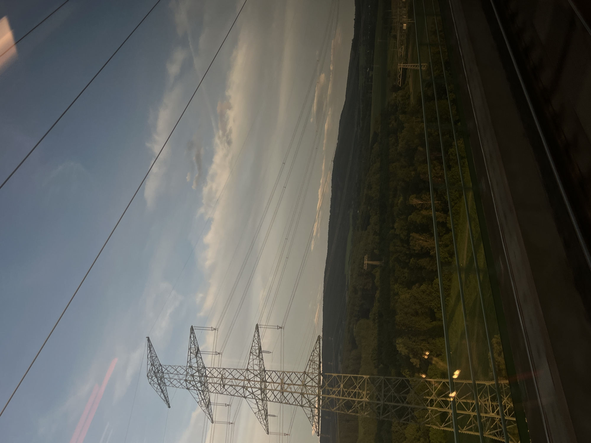 A vast, green landscape unfolds under a sky of scattered clouds. In the foreground, a tall, metal power pylon stands prominently, its lattice structure reaching towards the heavens. Power lines stretch horizontally across the frame, converging towards the horizon. The lush vegetation suggests a rural or semi-rural setting, possibly a park or forest bordering a town. The scene appears to be captured from a moving vehicle, perhaps a train, given the slight blur and reflections on the window. The lighting indicates it is either early morning or late afternoon, with the sun casting a warm glow on the landscape. This power pylon is located on the edge of Ilmenau, Germany, marking the transition from the urban environment to the surrounding natural beauty.