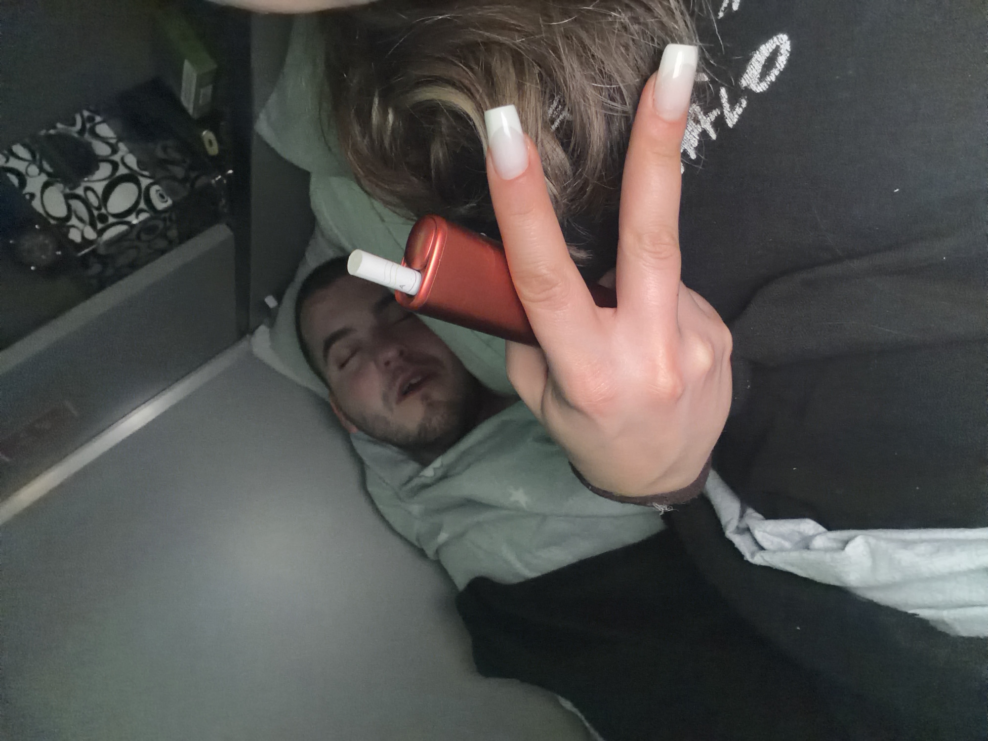 A person is asleep in bed, covered with a white blanket. A second person is above and holding a red vaping device with a white cigarette sticking out of it. They are holding up a peace sign over the head of the first person. There is a black t-shirt and light brown hair that belong to the second person. Beside the bed is a nightstand, a black and white design tissue box, and a green container. There is no explicit location given in this image to conclude it is in Varna, Bulgaria.