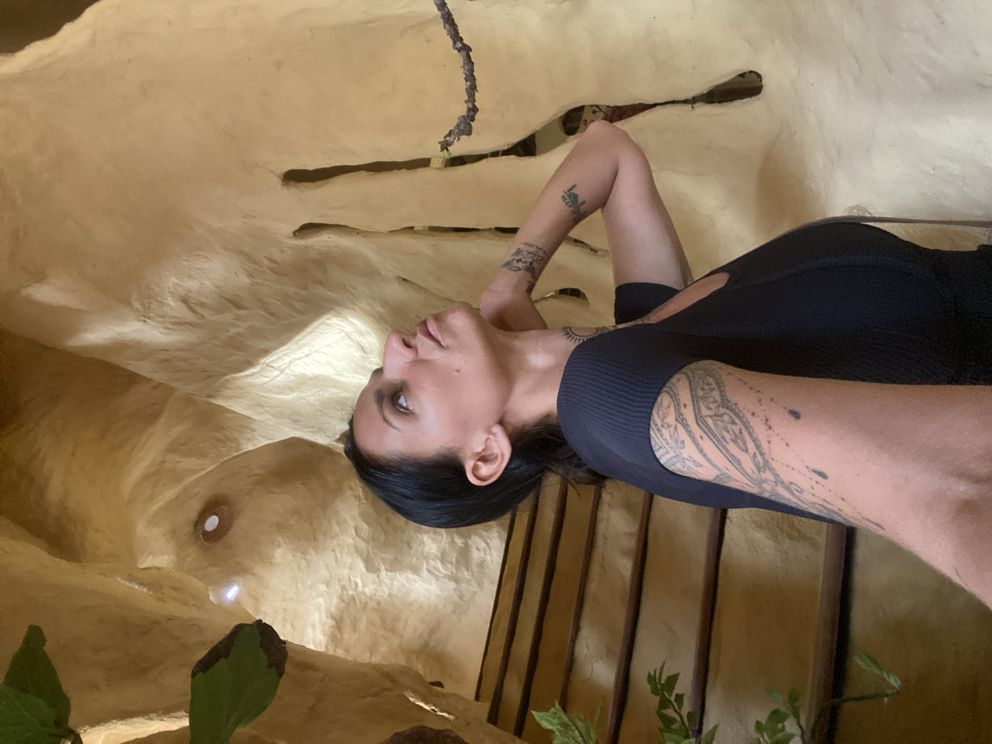 The image captures a woman with dark hair and tattoos on her arms, looking upwards within a cavernous interior. The walls are a textured, light yellowish-brown, resembling natural rock formations or sculpted earth. In the background, wooden stairs with dark railings ascend to the left, and lush green foliage is visible at the bottom left. To the right, the wall has elongated, irregular openings, and a thin, dry branch extends across the frame. The lighting appears to be artificial, with a small light source visible in the upper left portion of the cavern. This scene is located within the Fairy Stream (Suối Tiên) in Mũi Né, Vietnam, a popular tourist destination known for its unique geological formations and red sand dunes.
