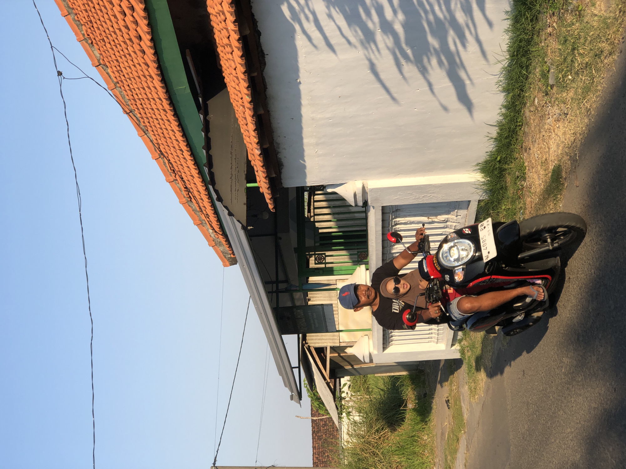 A man wearing a black t-shirt, blue baseball cap, and denim shorts rides a black scooter. He has a mustache and is looking forward. A young girl wearing a brown hijab and sunglasses sits behind him. She is wearing a red shirt. They are riding on a paved road in front of a white wall with a tiled roof. The sun is casting shadows on the wall. The background shows grass and trees. The license plate of the scooter is visible and reads "W 4085 PB". This scene is likely in Kalitengah, Indonesia, given the context provided.