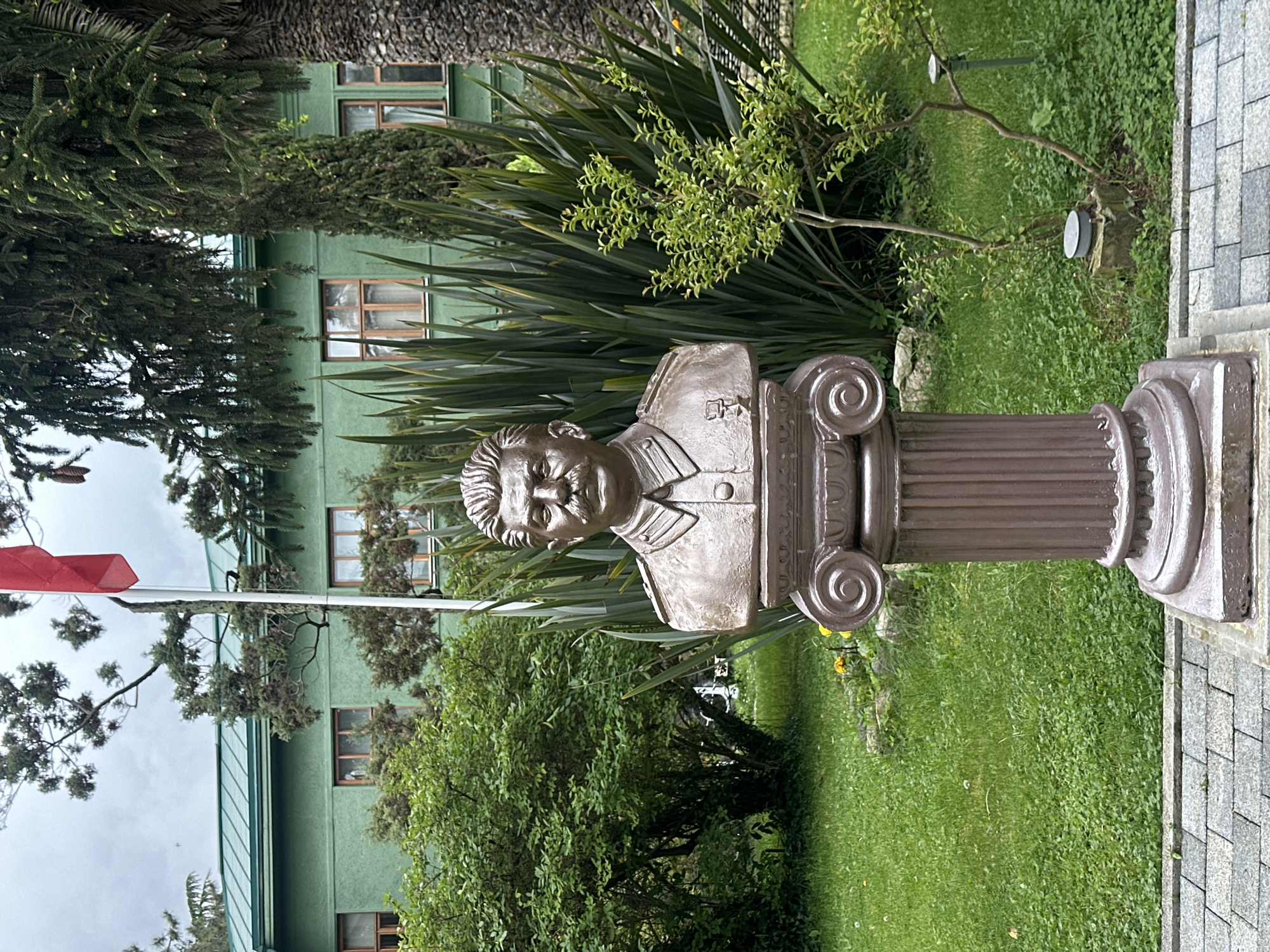 A bronze bust of Joseph Stalin stands on a fluted Doric column in an outdoor setting. The bust depicts Stalin in military attire, with a five-pointed star on his uniform. In the background is a green, multi-story building with evenly spaced windows. Surrounding the bust are various trees and shrubs, with a red flag visible in the background. The area adjacent to the column is paved with grey brick. This photograph was taken in Sochi, Russia.