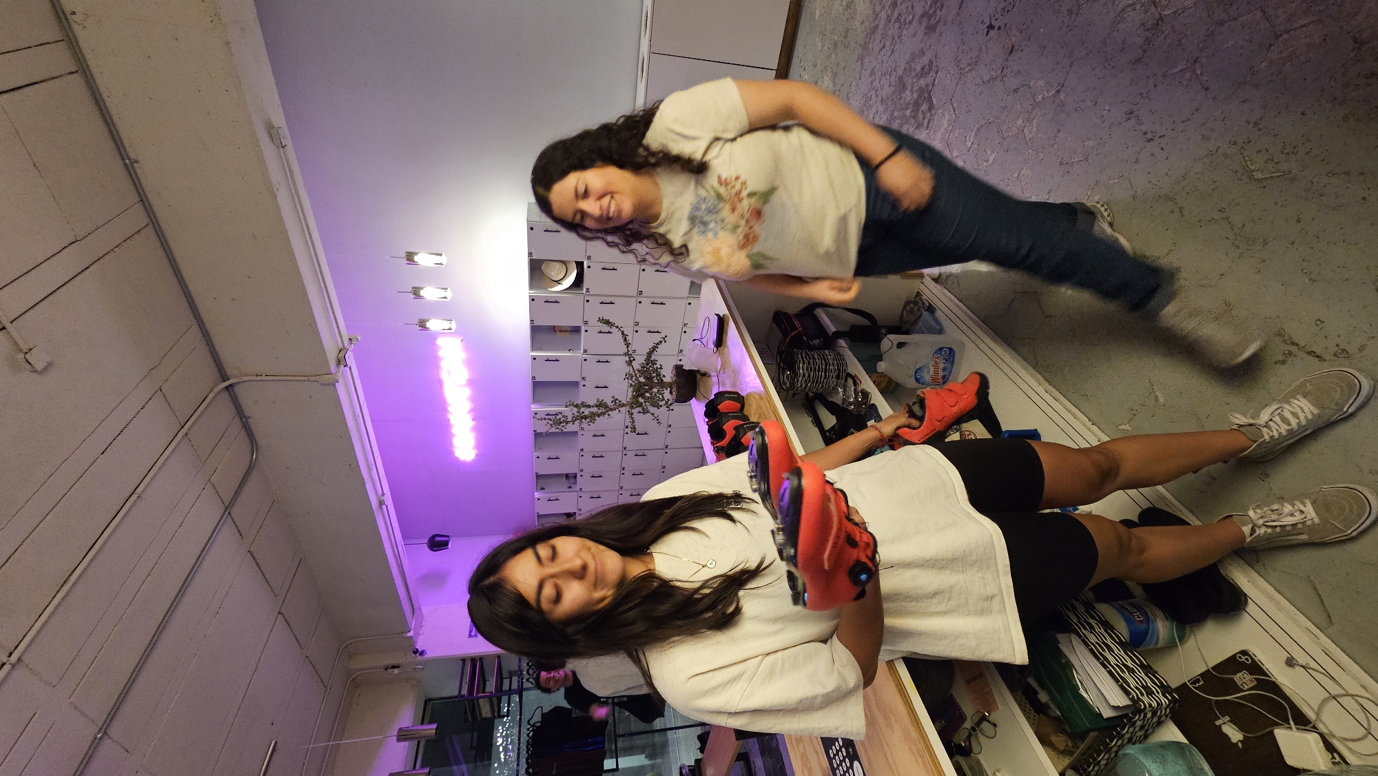 Two women are posing in what appears to be an indoor retail space or a storage facility, possibly in Puebla, Mexico. One woman is standing wearing a floral print t-shirt and jeans. The other woman is lying down on what appears to be a shelf unit, wearing a white t-shirt and dark shorts. She is holding a pair of red shoes. The background features locker-style storage units and a purple neon sign. Various objects and personal belongings are scattered along the shelf unit.