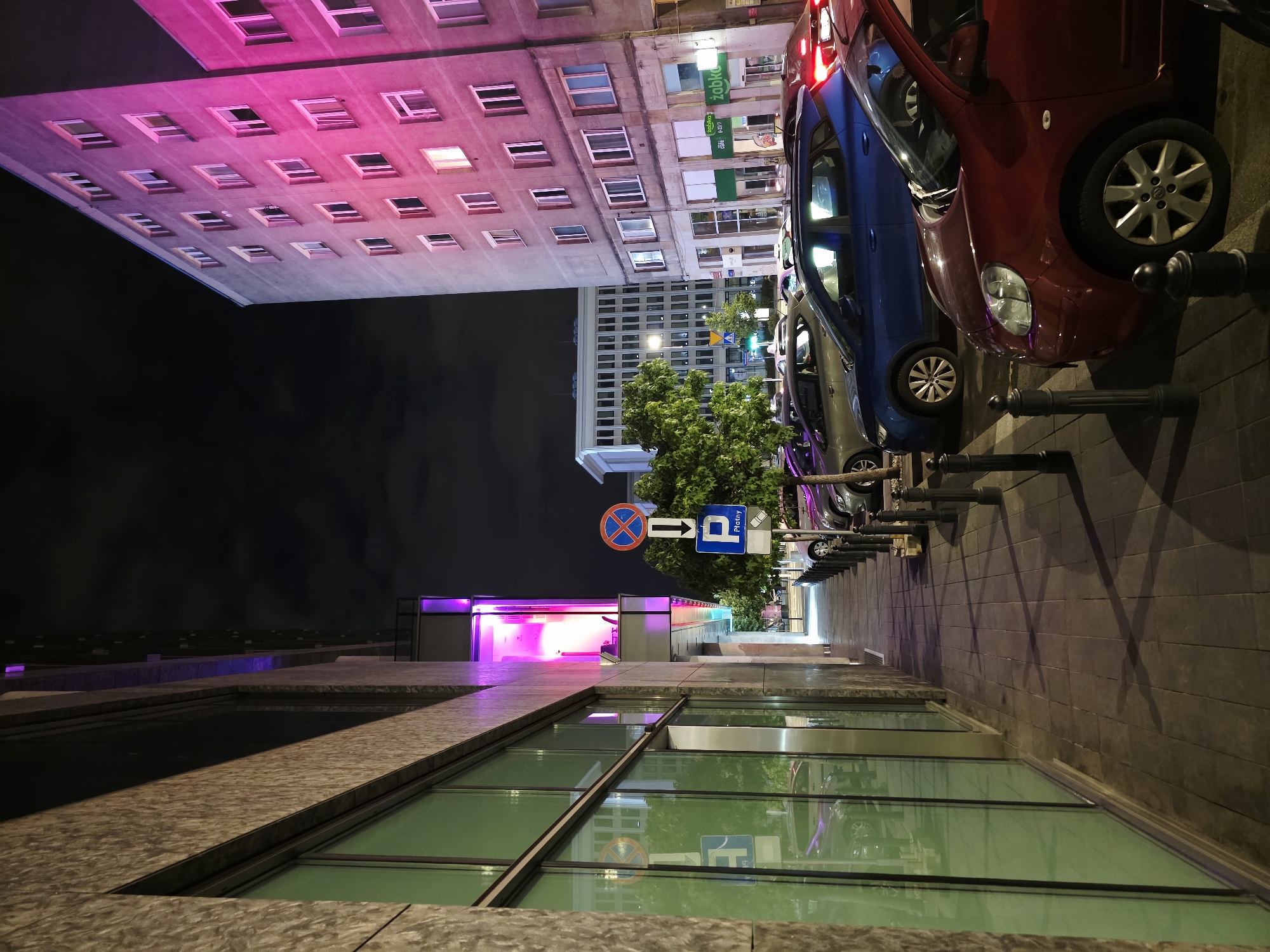 An urban street scene at night in Warsaw, Poland, features a sidewalk alongside a modern building with large green-tinted windows, illuminated by purple lighting from within. Across the street, a multi-story residential building is brightly lit with pink and purple lights, highlighting its numerous windows. Several cars are parked along the sidewalk, including a red hatchback and a blue SUV. Traffic signs, including a parking sign with an arrow, are visible. The sky is dark and cloudy. The scene captures a moment of urban night life with architectural lighting and parked vehicles.