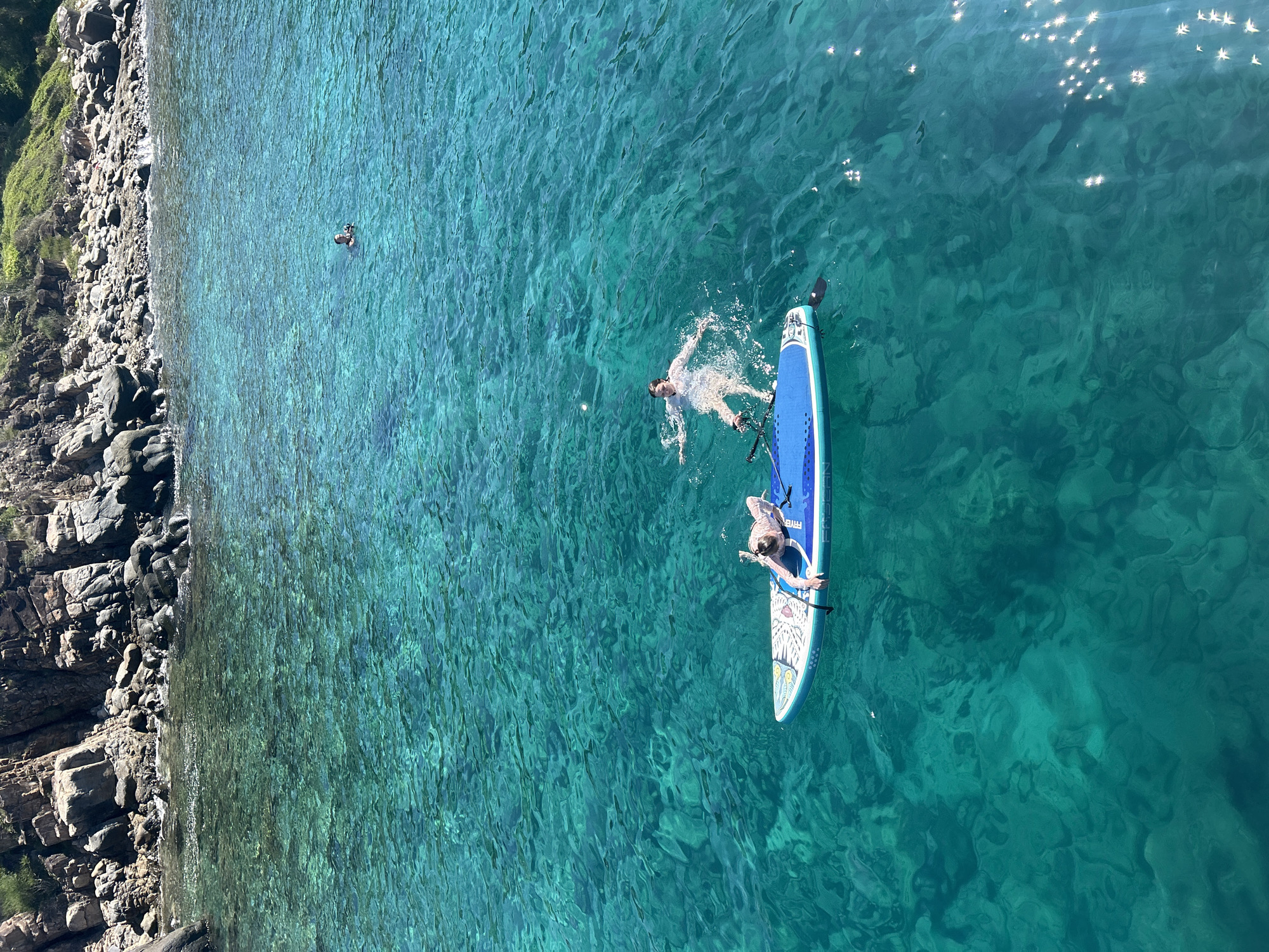 The image depicts a scene in the clear, turquoise waters off the coast of Hon Mun Island, within Nha Trang Bay, Nha Trang Ward, Vietnam. In the foreground, a blue stand-up paddleboard floats on the water. Two individuals are present. One person is lying on their back on the paddleboard, with their arms extended. Another person is in the water next to the paddleboard, with their body splayed out and arms outstretched. Further in the background, a third individual is swimming in the water, with their head visible above the surface. The background features a rocky, vegetated coastline. The water is shallow enough to see the seabed beneath, with sunlight creating shimmering patterns on the surface.