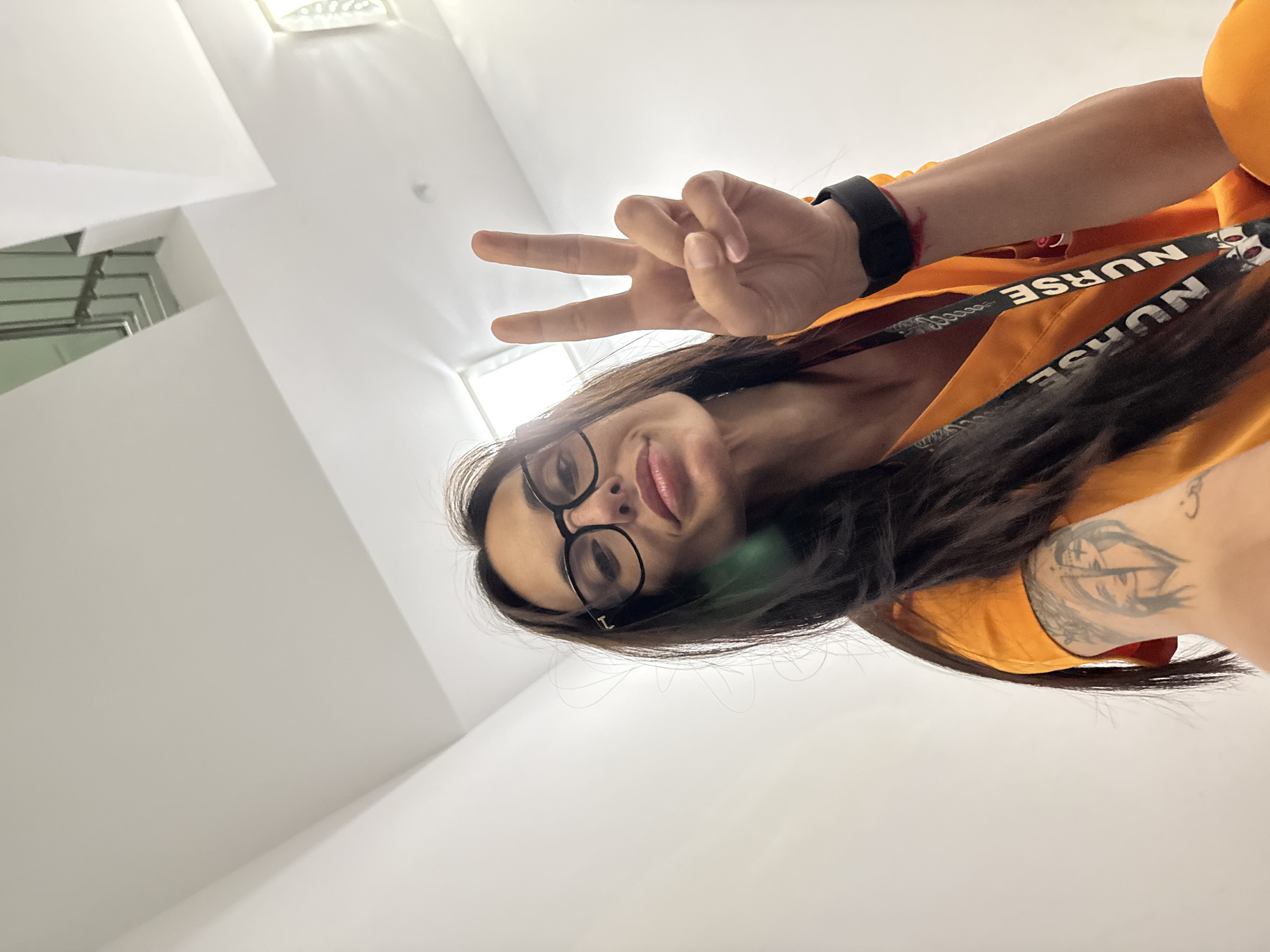 A low-angle, tilted selfie shows a woman with long dark hair and glasses, making a peace sign. She is wearing an orange top and a lanyard with the word "NURSE" printed on it. A tattoo is visible on her arm. The background is a plain white ceiling with a light fixture. There is no information in the image to indicate a location in Pleven, Bulgaria.