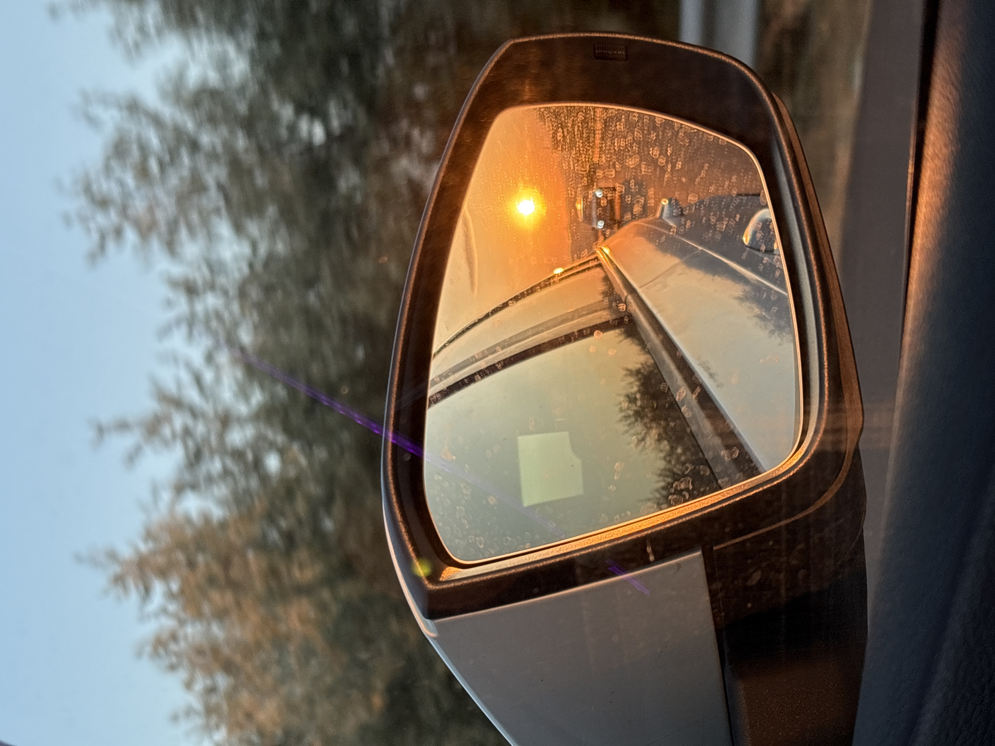 A side view mirror of a vehicle reflects a sunset over a road. The mirror is angled to show the setting sun, which is a bright orange orb. The sky is a clear blue, and trees line the background. In the reflection, other cars can be seen on the road, their headlights illuminated. The mirror itself is slightly dirty, with water droplets and streaks visible on its surface. The setting is consistent with a rural area, possibly on the outskirts of a town like Bulgarovo, Bulgaria, where such scenic drives are common.