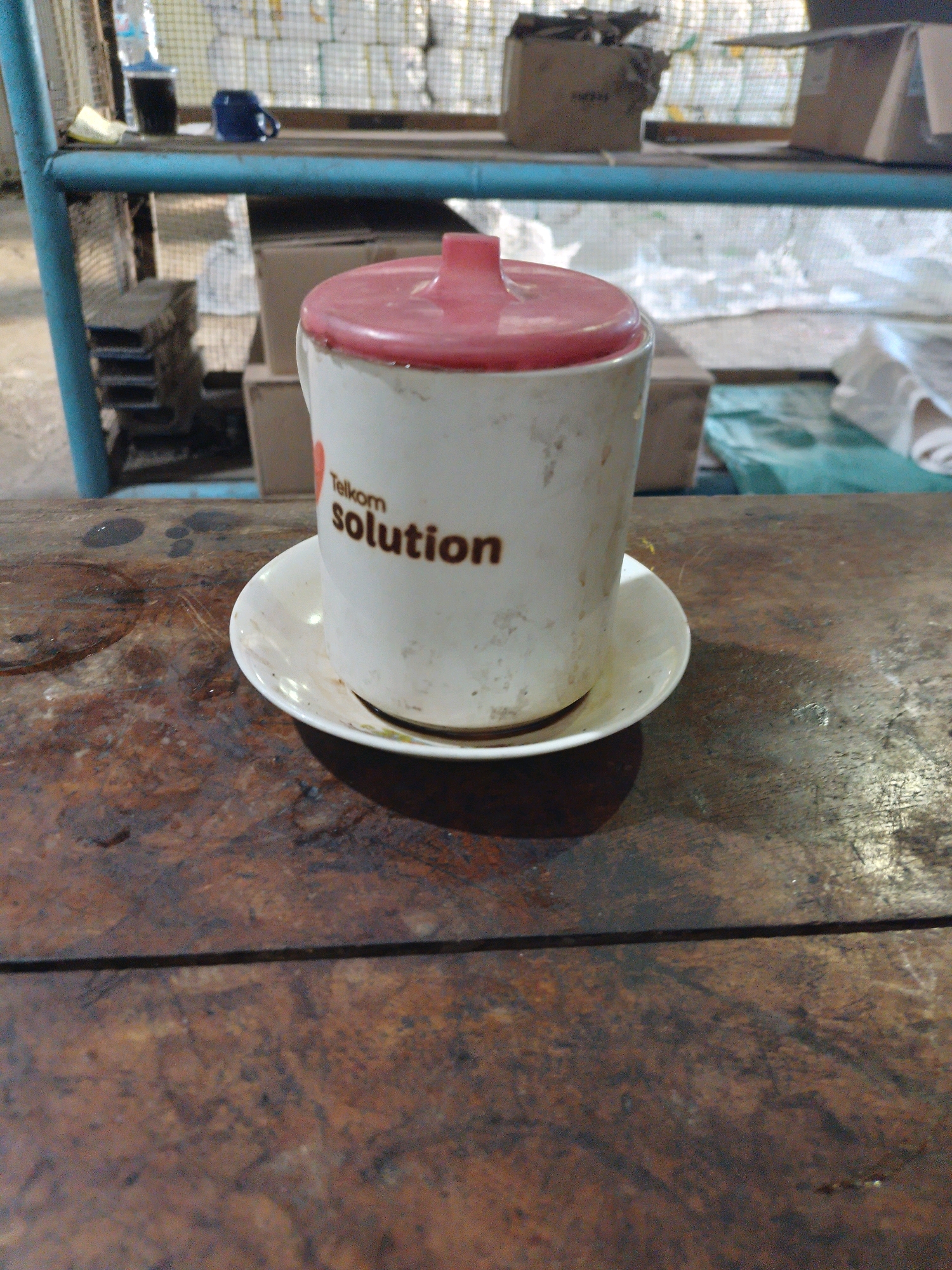 A white mug with a red lid sits on a white saucer. The mug has the words "Telkom solution" printed on it in brown lettering. The mug and saucer are dirty and appear to have residue on them. The mug and saucer are placed on a dark, rustic wooden surface. In the background, a metal shelf is visible, with various objects on it, including a blue mug, a bottle of water, and cardboard boxes. The overall environment appears to be utilitarian or storage-like. The location is Patianrowo, Indonesia.