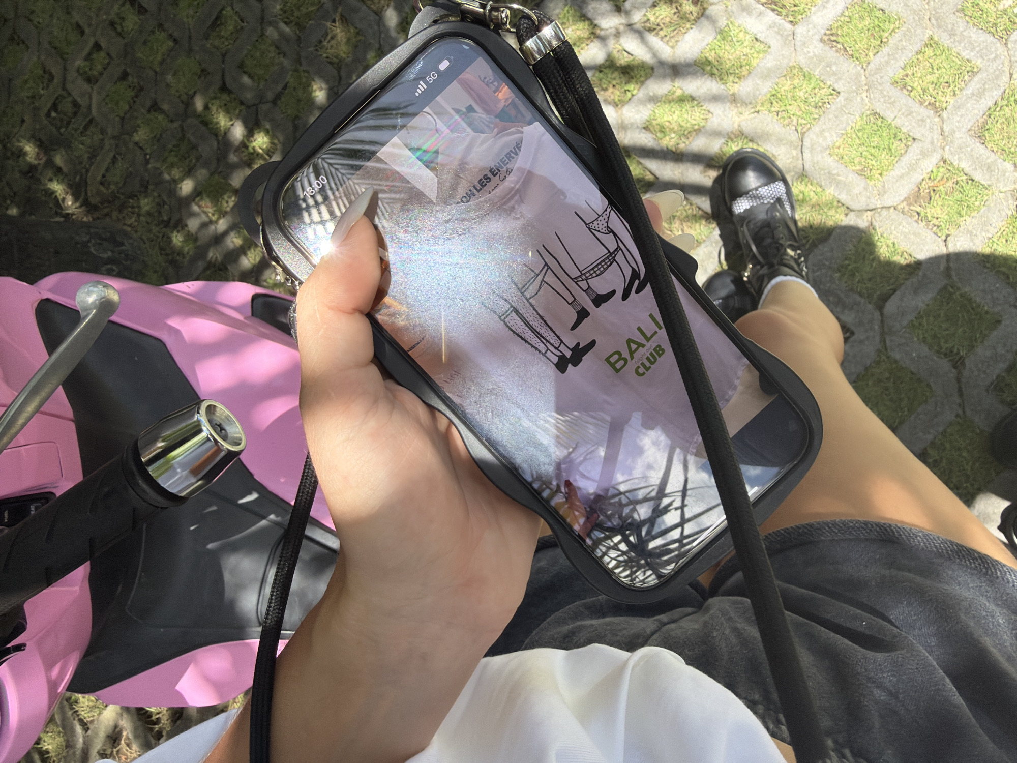 A first-person, close-up shot captures a hand holding a smartphone with a black case and neck strap, displaying an image of an advertisement for "Bali Night Club," with cartoon illustrations of figures in minimal clothing. The phone is being held above a pink scooter in Canggu, Indonesia, based on the location advertised on the phone's screen. A portion of the scooter's black handlebar and chrome end is visible. The person is wearing grey denim shorts, black boots, and a white shirt. The background features a paved surface with grass growing between the stones.