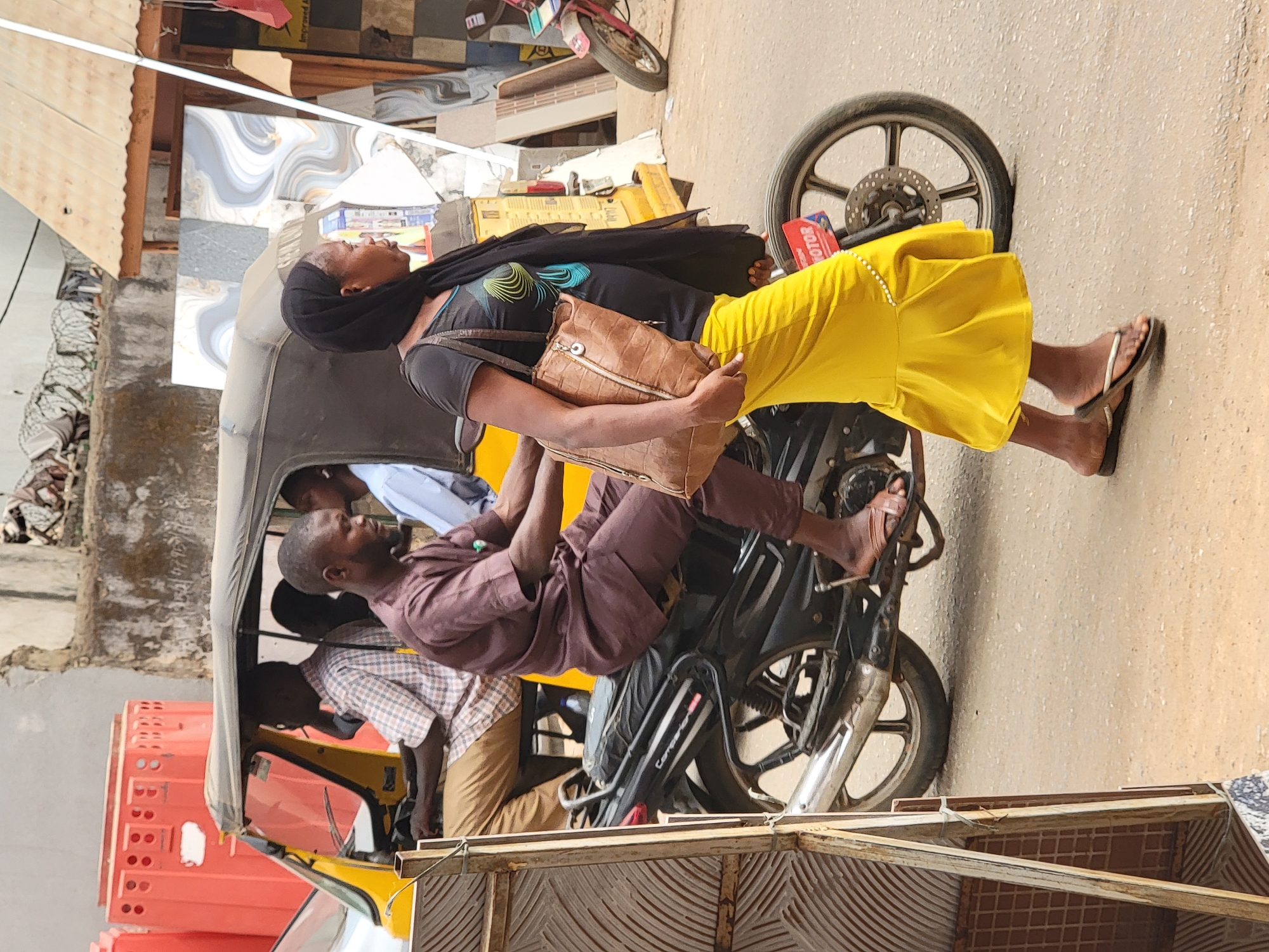 The image depicts a scene in Maiburiji, Nigeria. A woman wearing a black headscarf, a grey shirt, and a yellow skirt is seated behind a male passenger on a motorcycle taxi. The male passenger is wearing a brown shirt and pants. The motorcycle is part of a yellow three-wheeled vehicle with a canopy. Another man is seated in the front of the vehicle. The background shows buildings and street details.