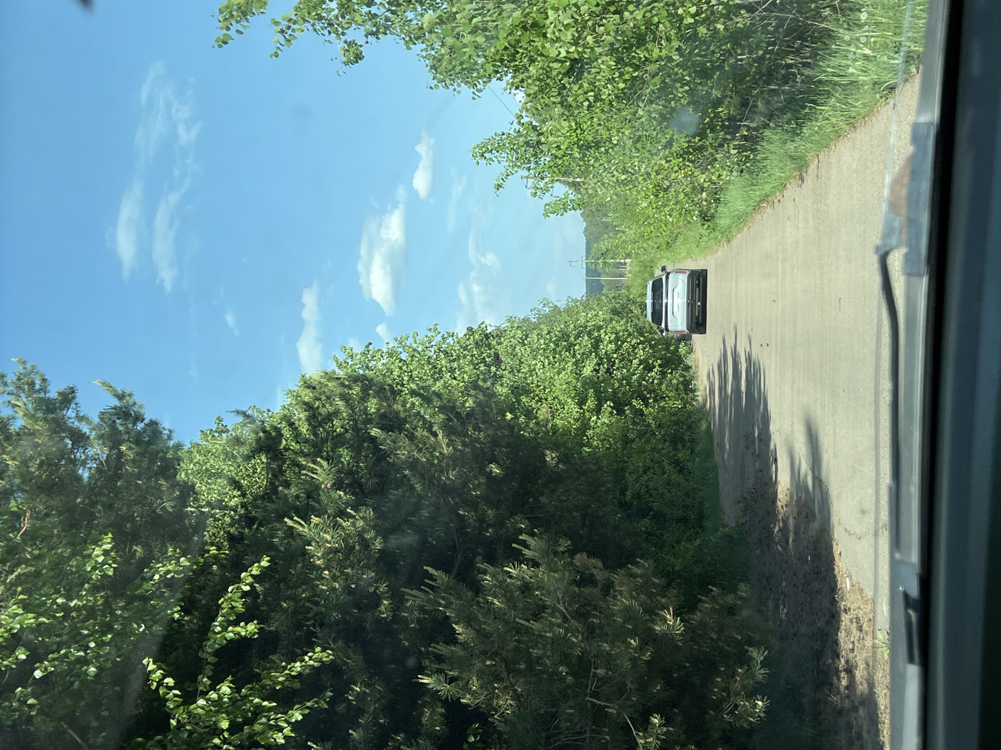 A view from inside a vehicle looks out onto a narrow, paved road lined with lush green vegetation on both sides. A white or light-colored van is driving ahead on the road. The sky is blue with scattered white clouds. The scene appears to be a rural or suburban area, possibly on the outskirts of a city. It is not possible to identify the location as Chaikovsky, Russia, from the image alone. There are no visible people or interactions.