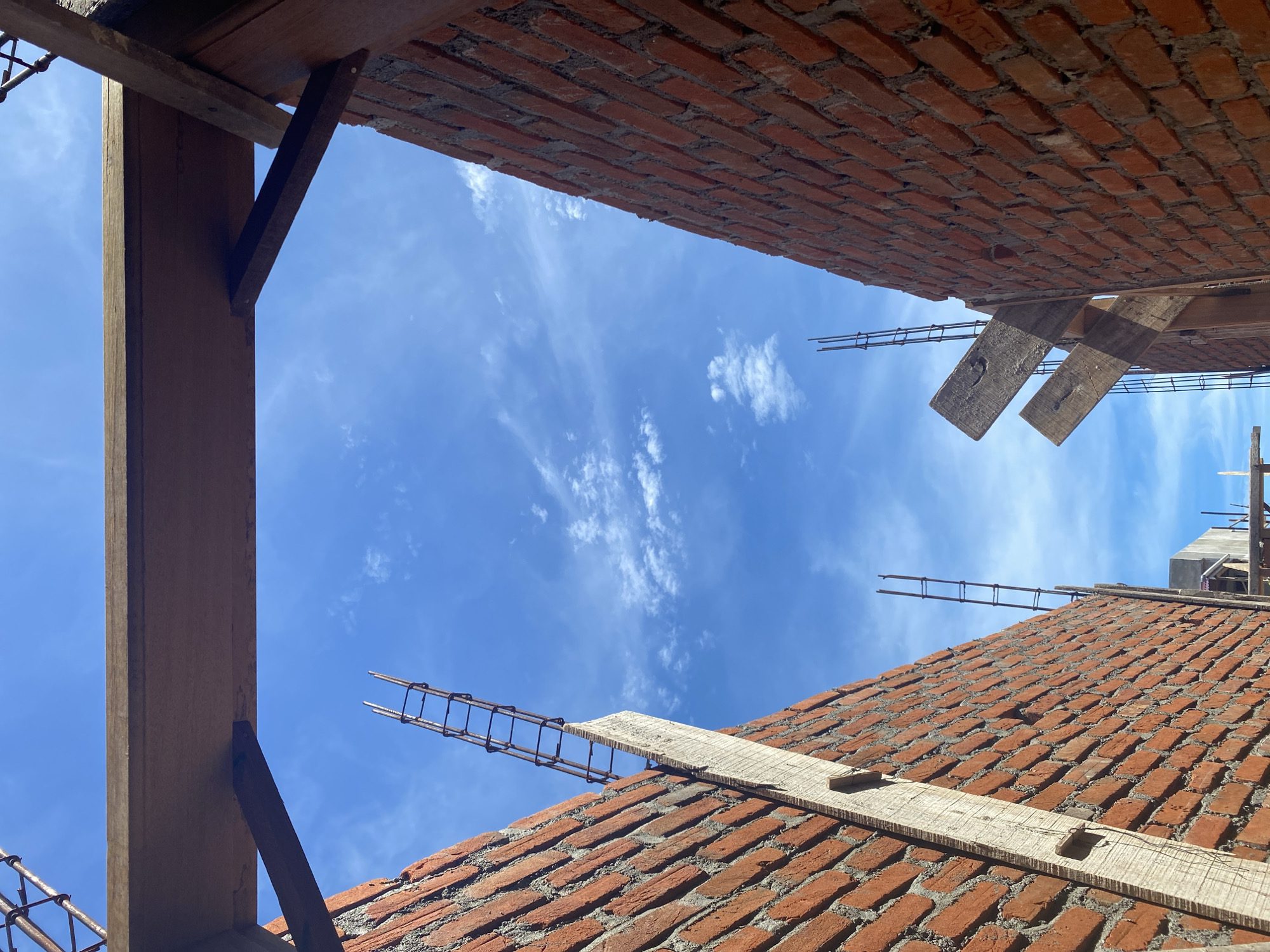 The image depicts a construction site under a bright blue sky with wispy clouds. Two brick walls, constructed from reddish-orange bricks with grey mortar, run parallel to each other, creating a narrow passageway. Rebar extends vertically from the top of the walls, indicating ongoing construction. Wooden beams and supports are also visible, some bridging the gap between the walls and others attached to the brickwork. The perspective is looking upwards, emphasizing the sky and the unfinished structure. The location is identified as Banda Aceh, Indonesia.