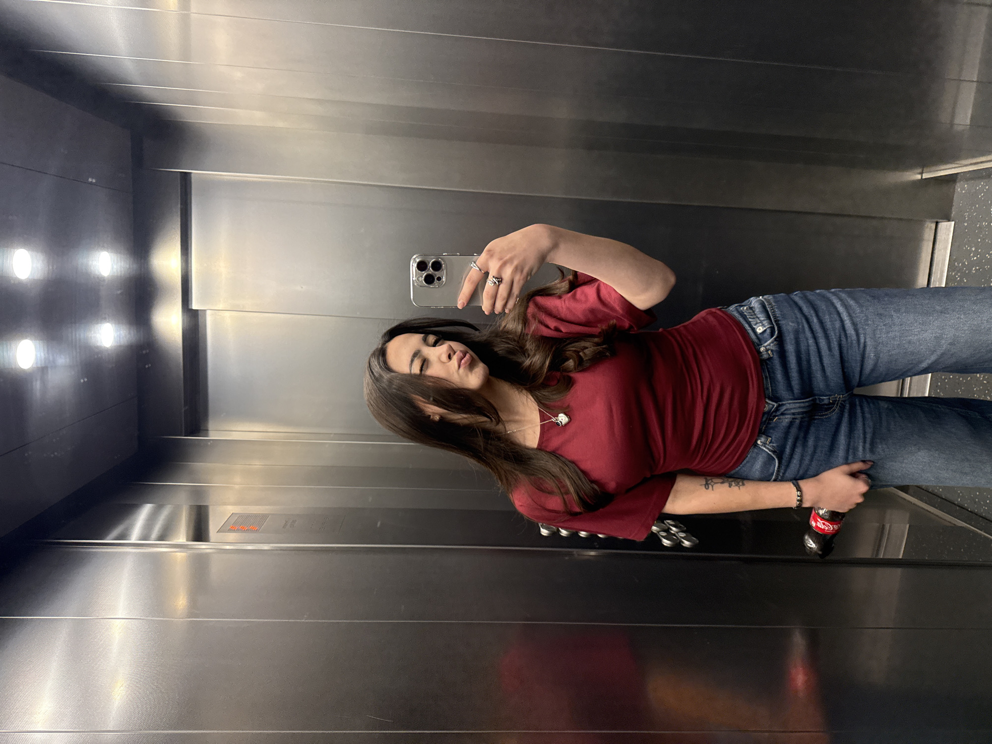 A woman is taking a selfie in an elevator. She is wearing a red long-sleeved shirt and blue jeans. The elevator has stainless steel walls and ceiling. There is a bottle of Coca-Cola in her hand. The location is not explicitly stated as Yerevan, Armenia, and cannot be confirmed from the image.