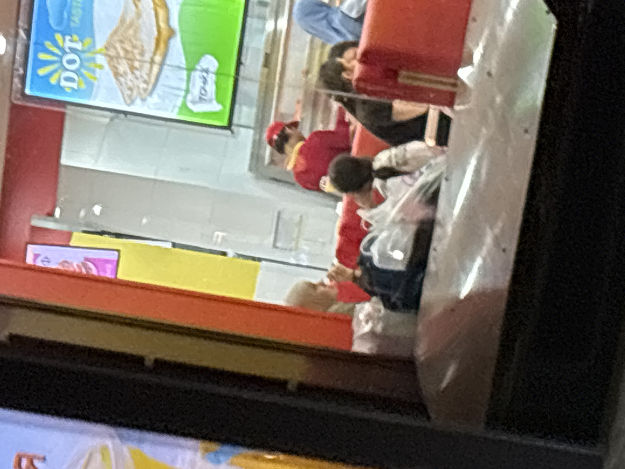 The image shows a blurry scene inside a food establishment, possibly a fast-food restaurant or cafe, viewed through a window. The interior features bright, colorful signage, including a sign with yellow sunburst graphics and the word "DOT" above an image of food. There are several people present, some appear to be customers and at least one might be an employee wearing a red cap. They are seated or standing behind a counter area. The overall impression is of a casual dining environment, but the low image quality and the viewpoint through glass make precise identification of objects and activities difficult. There is no information within the image or its crops that would allow for identification of the location as Grozny, Russia.