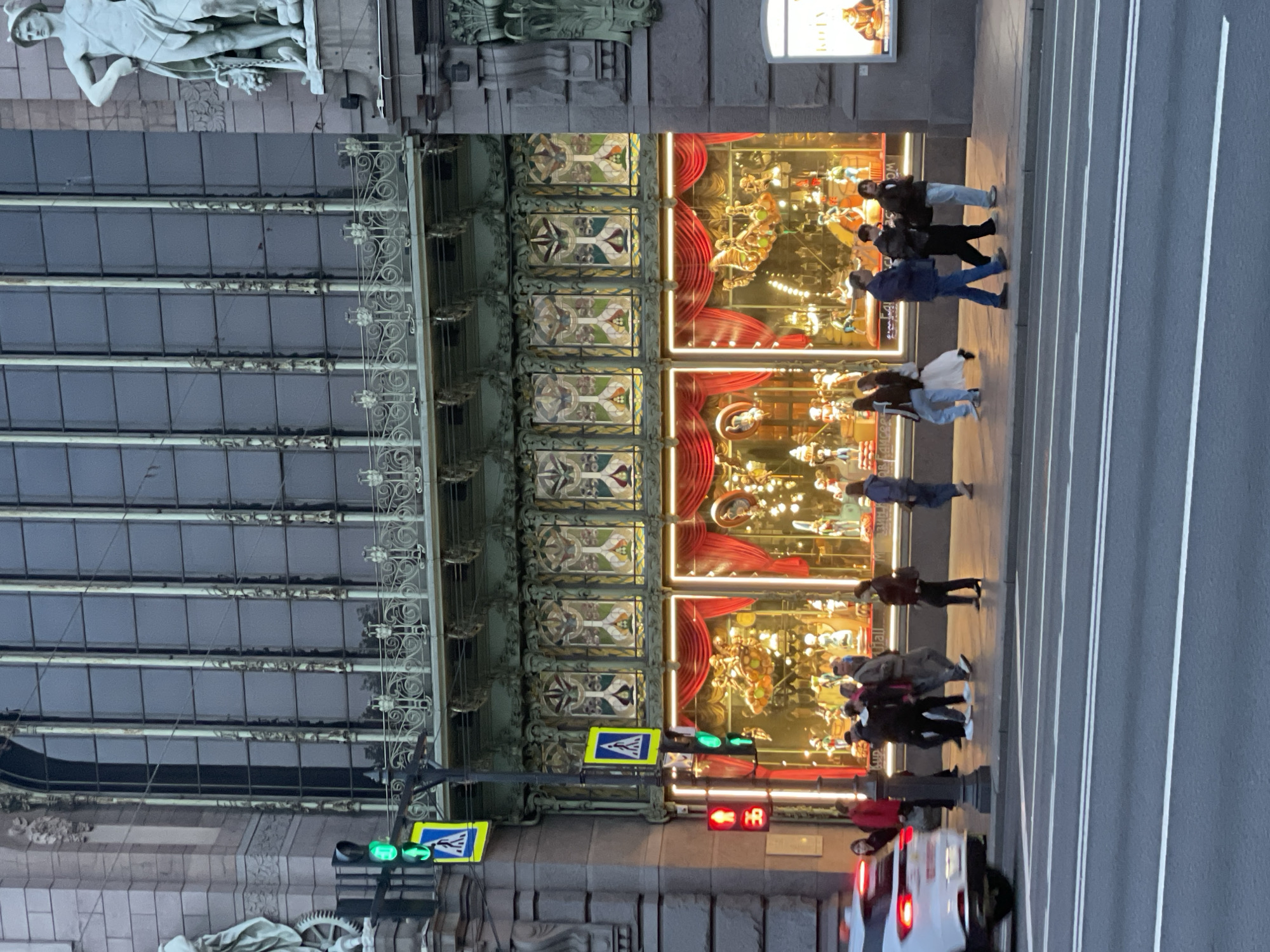 The image captures a street scene in Saint Petersburg, Russia, identifiable by the distinctive architectural style and landmarks. Focus lies on the intersection and adjacent building facades. Pedestrians are observed crossing the street. There is a white car is turning a corner. Traffic lights are present, with signals displayed. The architectural details suggest a historical location within the city. The building facade showcases decorated windows with carousel displays. Sculptures are visible atop the building.