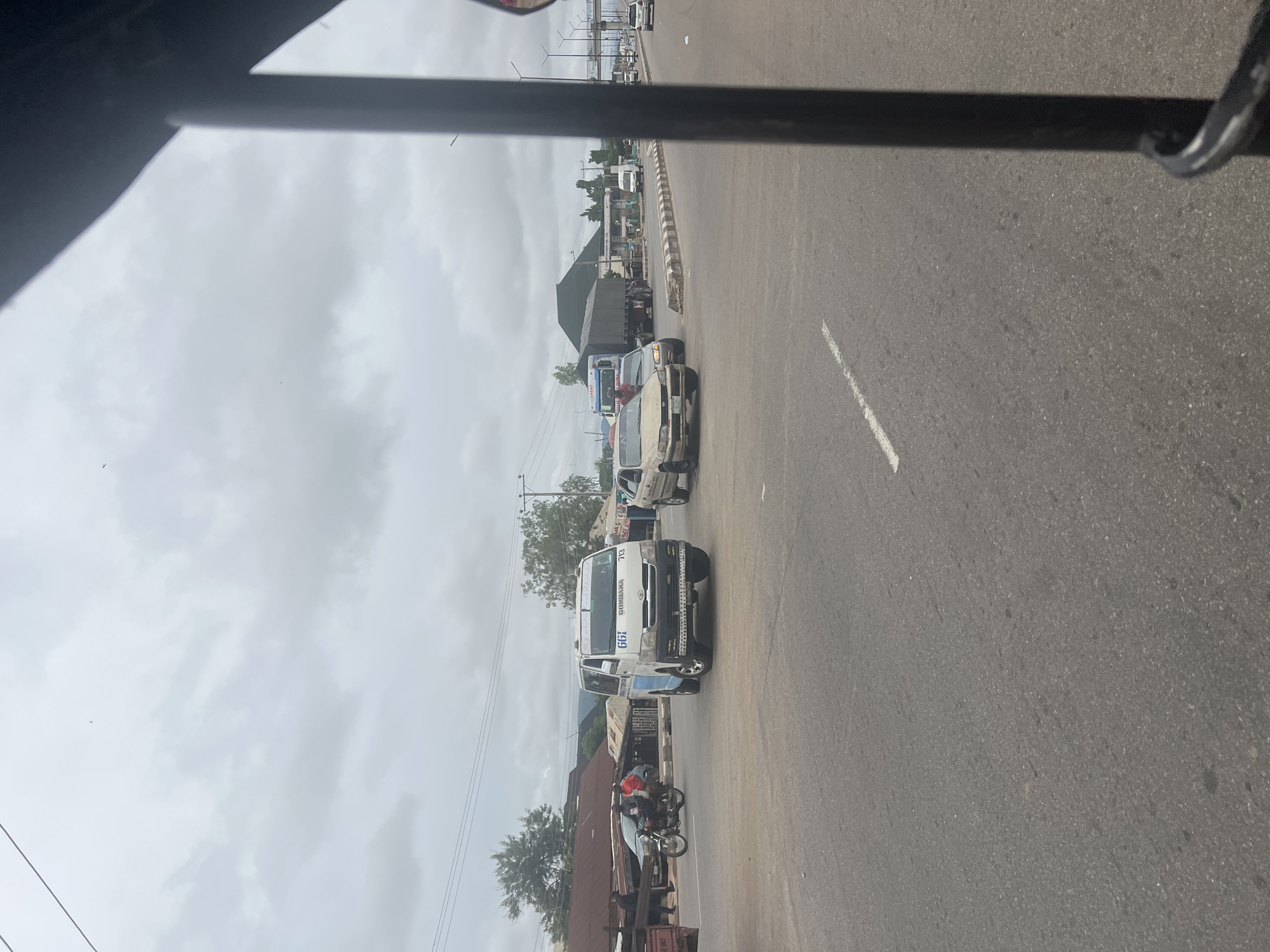 This is a street-level, eye-level, medium shot taken from inside a vehicle, looking out at an intersection in Jalingo, Nigeria. The sky is overcast with gray clouds. In the foreground, the asphalt road is visible with white lane markings. A black metal bar, likely part of the vehicle the photo is taken from, is on the right side of the frame.
At the intersection, a white minibus with blue accents and the text "GOMBARA" and "713" on its side is positioned in the middle lane, facing forward. To the left of the minibus, a motorcycle with two passengers is visible. Further left, a red object, possibly part of another vehicle, is partially seen. Behind the motorcycle, a row of buildings with corrugated metal roofs lines the street.
In the background, to the right of the minibus, a beige sedan is in the lane, and beyond it, a large articulated truck with a gray tarp covering its trailer is at the intersection. Several other vehicles, including a white van and cars, are further down the road. A pedestrian bridge is visible in the distance on the right. The environment appears to be an urban or semi-urban area with a mix of commercial and residential structures.