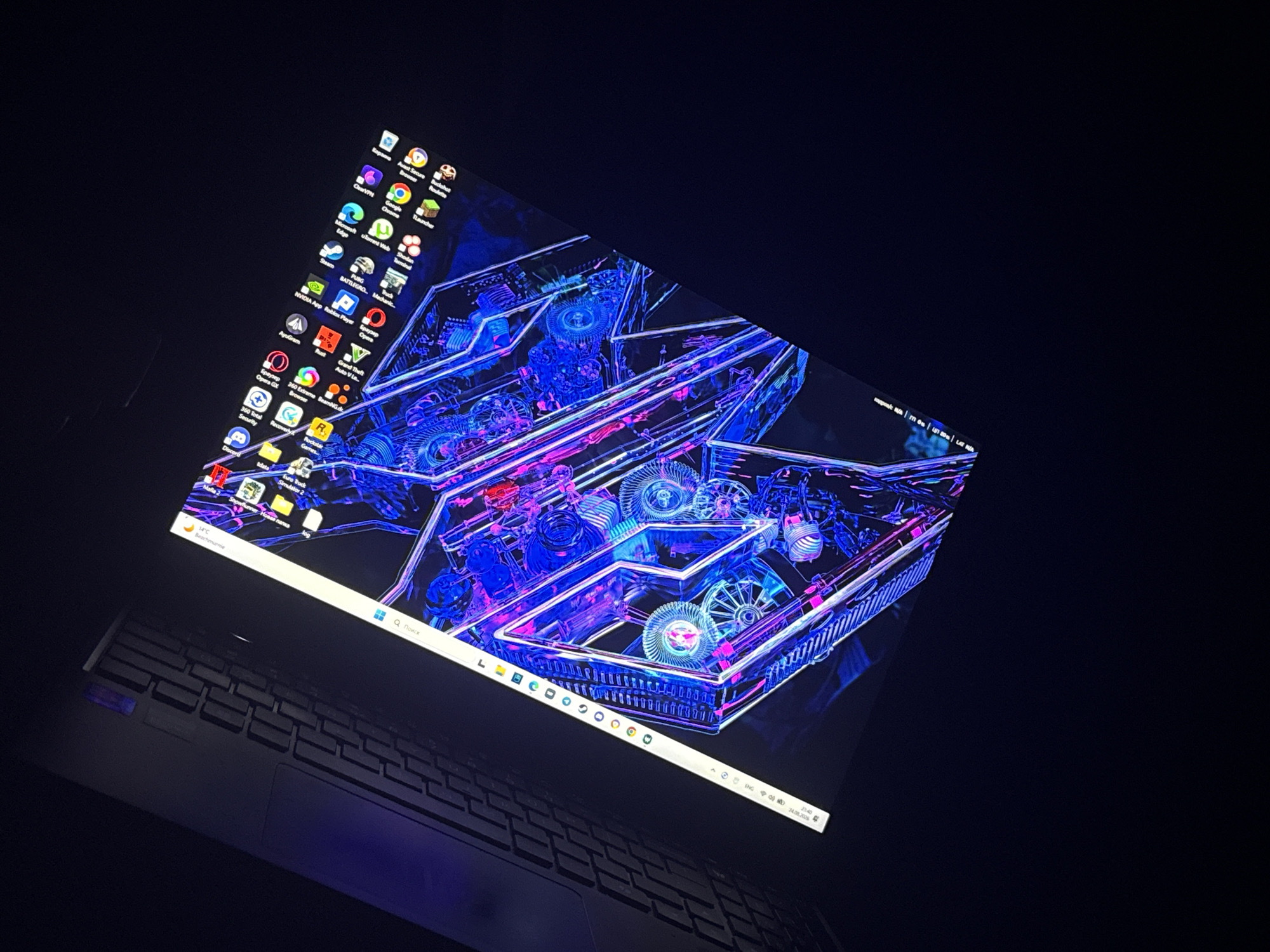 The image shows a laptop with its screen displaying a colorful, abstract geometric wallpaper with a blue and pink neon aesthetic. The laptop is angled in the dark, with only the screen and a portion of the keyboard illuminated. Icons are visible on the left side of the screen, suggesting a Windows operating system. The taskbar at the bottom of the screen shows various application icons, including what appears to be a search bar, a Windows logo, and several browser icons. The bottom right corner of the screen displays system information such as the time and date, reading "21:40 24.08.2023". The scene is indoors, with the ambient lighting primarily coming from the laptop screen itself. There is no indication of the city of Ustronie Morskie, Poland, or any specific outdoor environment. The image is focused on the laptop and its display.