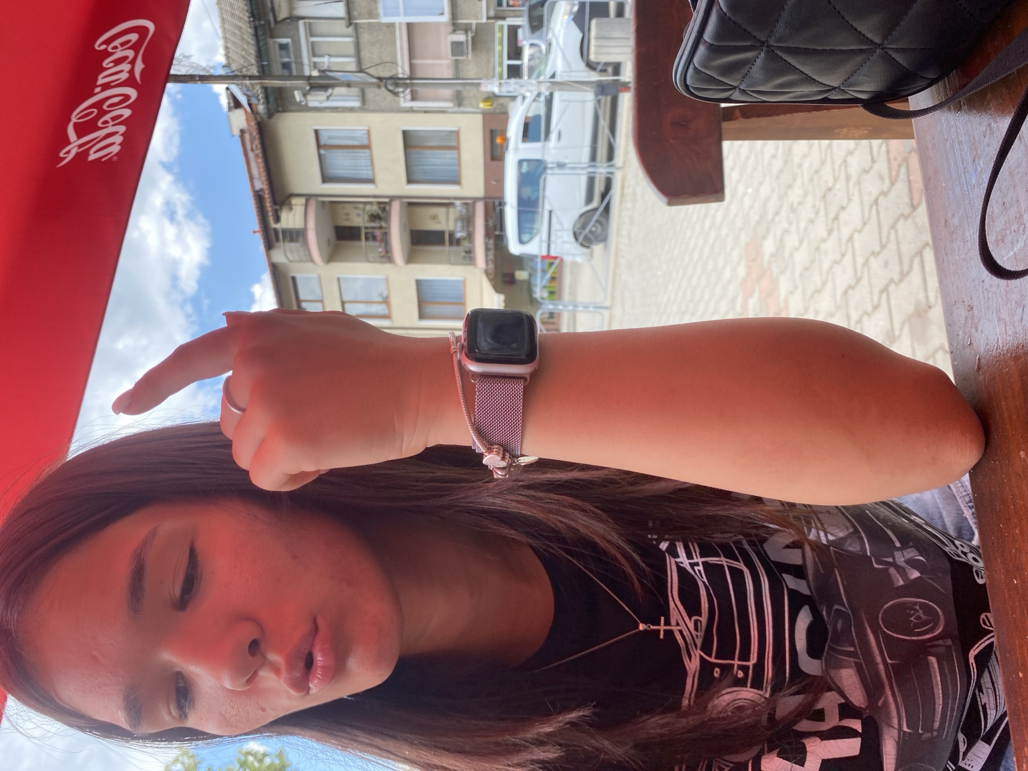 A young female with long, dark hair is captured in a close-up shot, leaning forward with her lips pursed and her right index finger extended upwards, as if making a point. She wears a black t-shirt with a graphic design featuring cars and the word "RACING," along with a silver cross pendant necklace. On her wrist, she sports a pink mesh Apple Watch band and a charm bracelet with a light-colored stone and a small silver dangle. The background reveals a street scene in Sevlievo, Bulgaria, with buildings, a parked white van, and a Coca-Cola branded umbrella overhead. A black quilted handbag rests on a wooden table in the foreground.