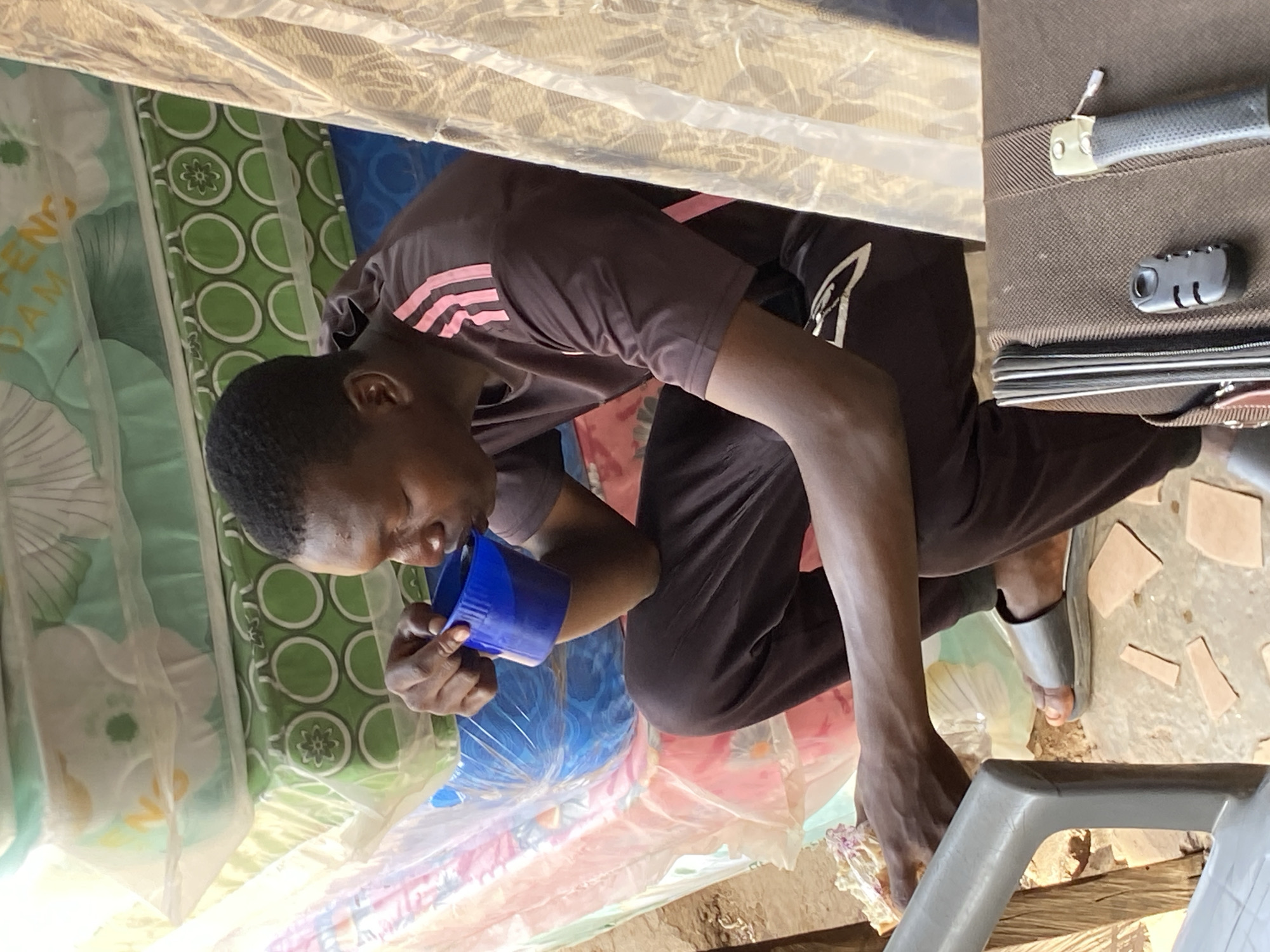 A man wearing a dark brown shirt with pink stripes is drinking from a blue cup. He is sitting in a narrow space, possibly inside a building in Bukuru, Nigeria, indicated by the presence of a mattress wrapped in patterned fabric on the left. The space is confined by the mattress, a beige fabric above, and luggage to the right with a combination lock. The floor is dirt and debris. The man is wearing gray sandals.