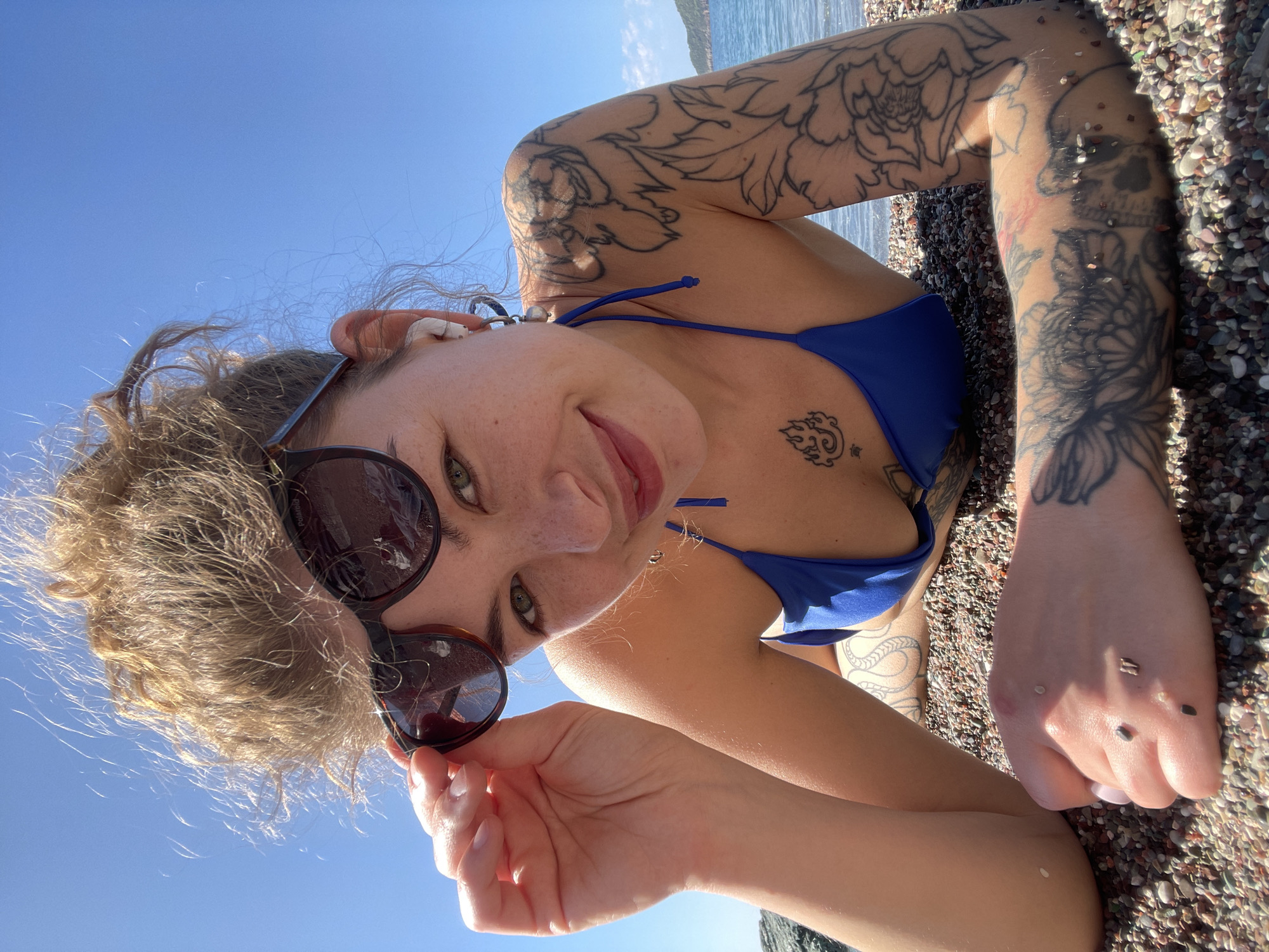 A close-up, rotated upward-facing shot captures a person, likely a woman, lying on a pebble beach in Rafailovići, Montenegro, under a clear blue sky. She is wearing a blue bikini top and bottom and looking directly at the camera with a slight smile. Her curly, light brown hair is pulled back, and she has green eyes. Sunglasses are perched on her head, with one arm of the glasses held by her hand, revealing "Polaroid" text on the frame.
Extensive tattoos adorn her arms and torso. Floral designs cover much of her left arm, while her right arm features similar patterns along with what appears to be a skull. A small tattoo resembling a cloud or flame symbol with a character is visible on her chest, and another tattoo is on her hip/side. Small, dark and light-colored pebbles are visible stuck to her skin, particularly on her arms and hands, indicating she has been in contact with the beach surface. The bright sunlight suggests it is daytime, creating strong shadows and highlighting the texture of the pebbles. In the background, the sparkling blue sea can be seen.