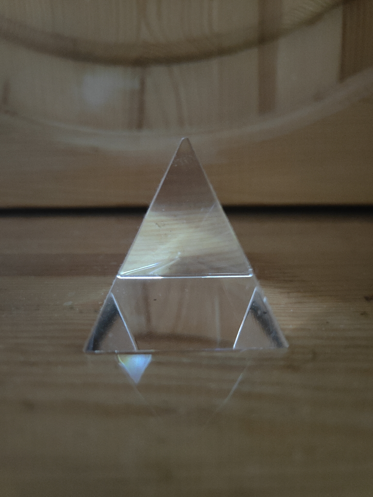 An upright triangular prism sits centered on a wooden surface. The prism is clear and reflects light, casting a faint rainbow-colored triangle onto the surface below. The wooden surface appears to be a tabletop or floor, with visible wood grain. In the background, a curved, lighter-colored surface is visible, possibly the inside of a bowl or container. The lighting is soft, creating subtle shadows and highlights on the prism and the wood.