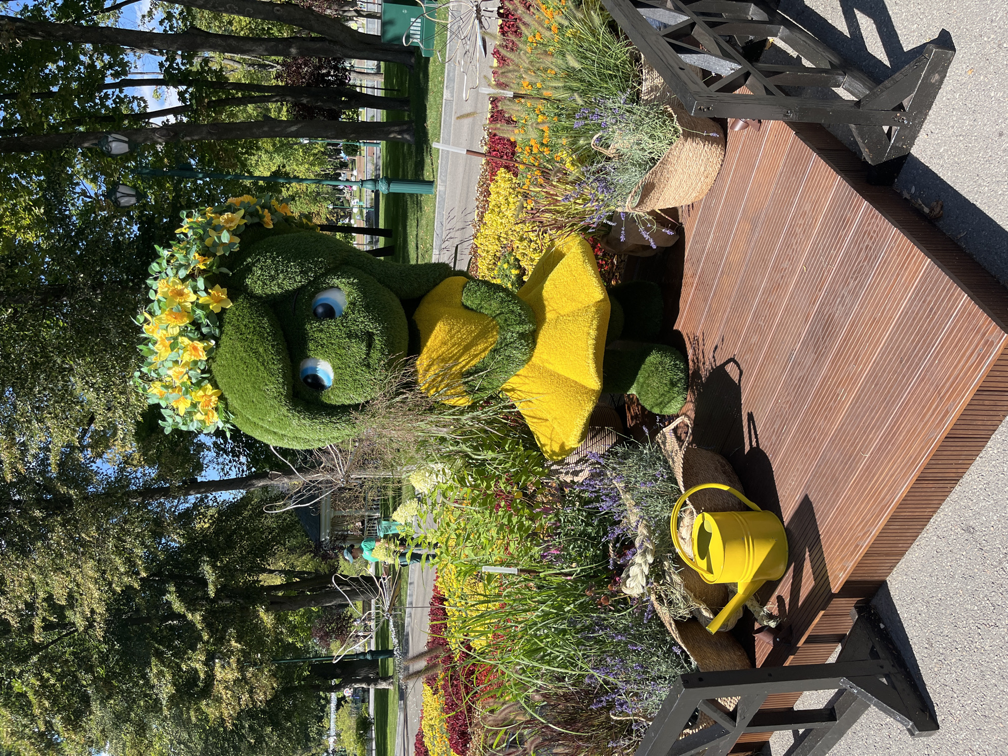The image depicts an outdoor scene in a park, likely within the city of Kharkiv, Ukraine, given the context of the provided reference material. The focal point is a large, green, topiary figure of a cartoonish character with large blue eyes and a yellow dress. The figure is adorned with a floral headdress featuring yellow flowers. It stands on a wooden platform in front of a colorful flowerbed filled with yellow, red, and purple blooms, as well as various grasses and lavender plants. To the left of the figure, on the wooden platform, sits a bright yellow watering can. In the background, trees, lampposts, and more flowerbeds are visible, suggesting a well-maintained public garden. A person in a green shirt and blue cap can be seen in the middle ground, appearing to be tending to the plants. The overall atmosphere is bright and summery, with sunlight casting shadows on the platform and the surrounding ground.