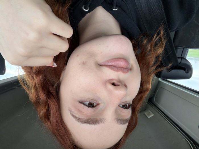 A young woman with auburn, wavy hair and light skin poses for a selfie in a vehicle. She wears a black hooded sweatshirt. Her left hand is raised near her face. The interior of the car is visible, with a rearview mirror and a gray ceiling or dashboard. The image is slightly angled.