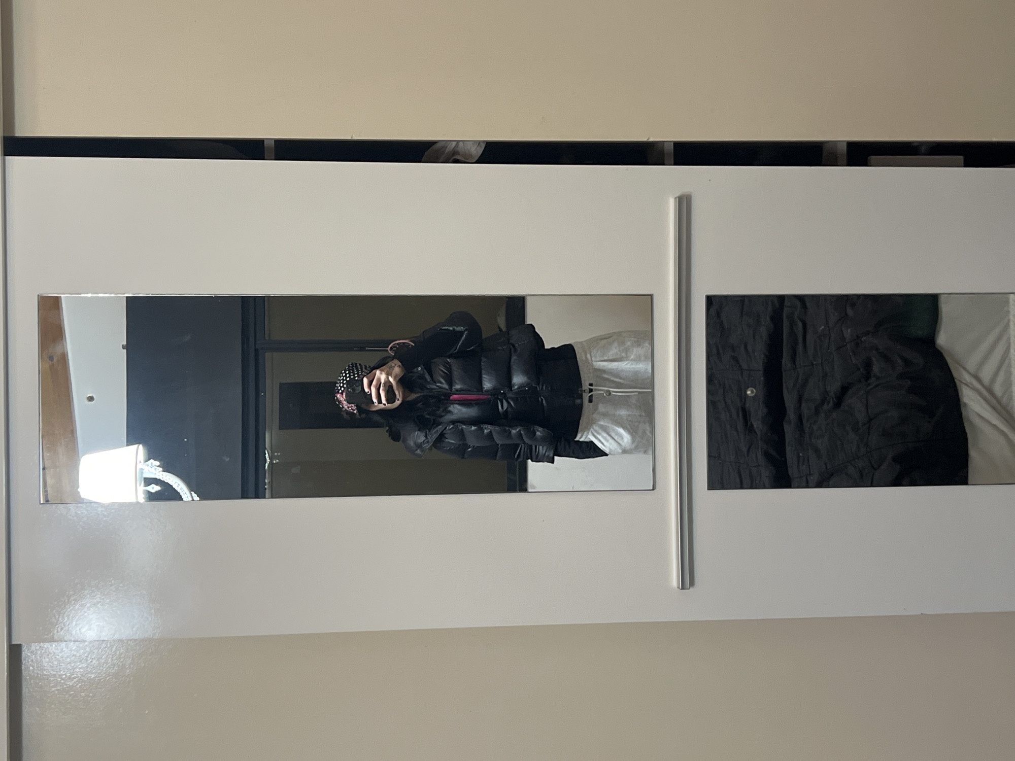 An indoor, vertically oriented image captures a mirror selfie taken by a person reflected in one of two mirrored panels integrated into white sliding doors. The person, positioned in the left mirror, is wearing a black puffer jacket, light-colored pants, and dark headwear with a white dotted pattern. They are holding a mobile phone to take the picture, partially obscuring their face. Their left hand shows some visible tattoos on the fingers and wrist.
The majority of the frame is dominated by the white closet doors, which have a thin silver vertical handle/divider between the two sections. The right mirrored panel reflects a dark, possibly quilted, fabric and a white surface, likely another piece of clothing or bedding. Above the white doors, there is a narrow black strip, and then a beige wall. A small gap is visible between the black strip and the beige wall, revealing a glimpse of an object behind the black strip. The reflection in the left mirror also shows a bright light fixture or object to the left of the person. There is no discernible text in the image. The time of day and specific location within Buenos Aires, Argentina, are not visually indicated beyond an indoor setting.