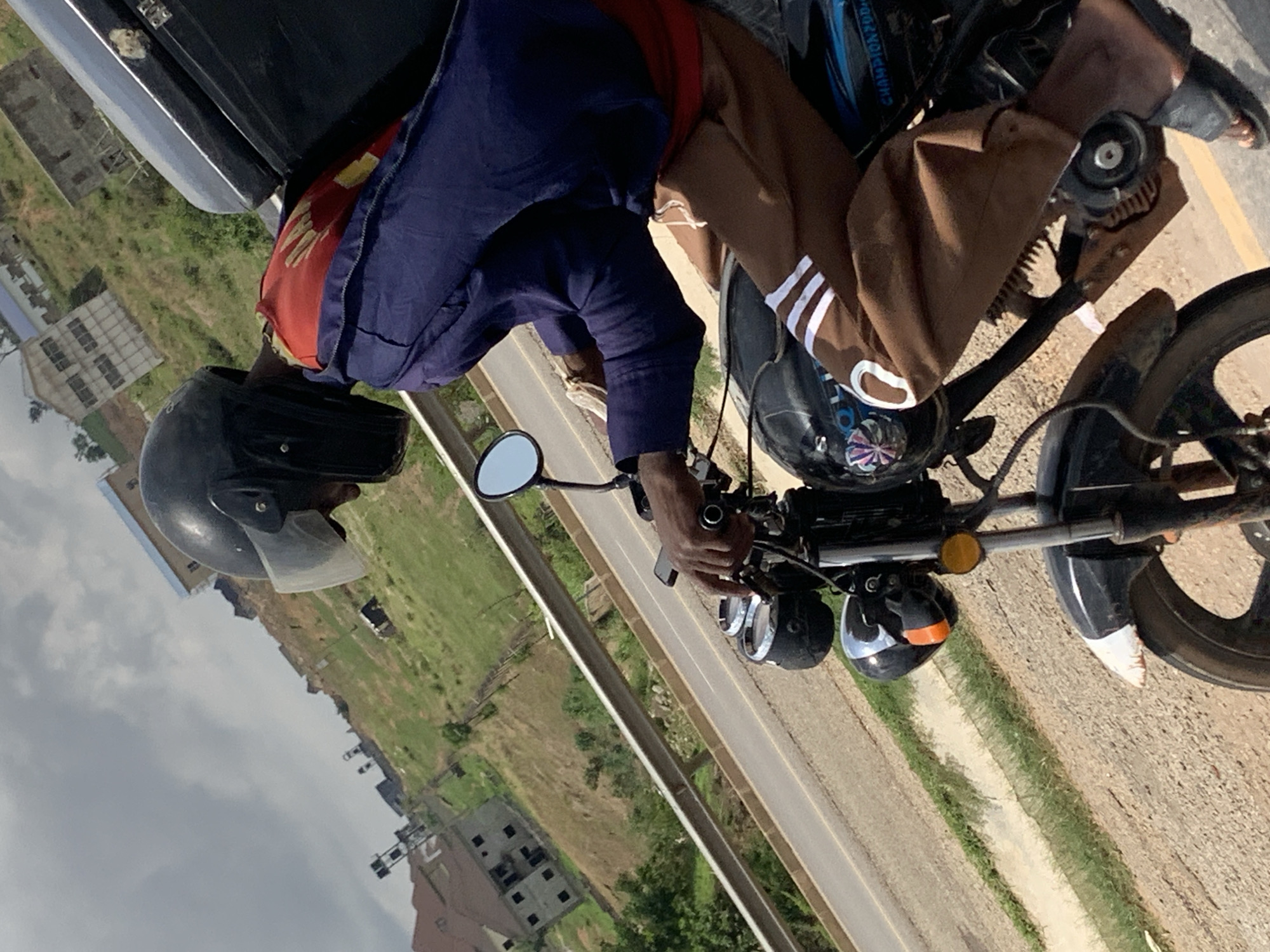 A man wearing a black helmet, a red shirt, a blue jacket, and brown pants with white stripes is riding a black motorcycle on a paved road. The road is lined with green vegetation and buildings in the background, suggesting a suburban or residential area. Based on the location, it's likely a street in Nyanya, Nigeria. The image is taken from a slightly elevated perspective. The day appears overcast.