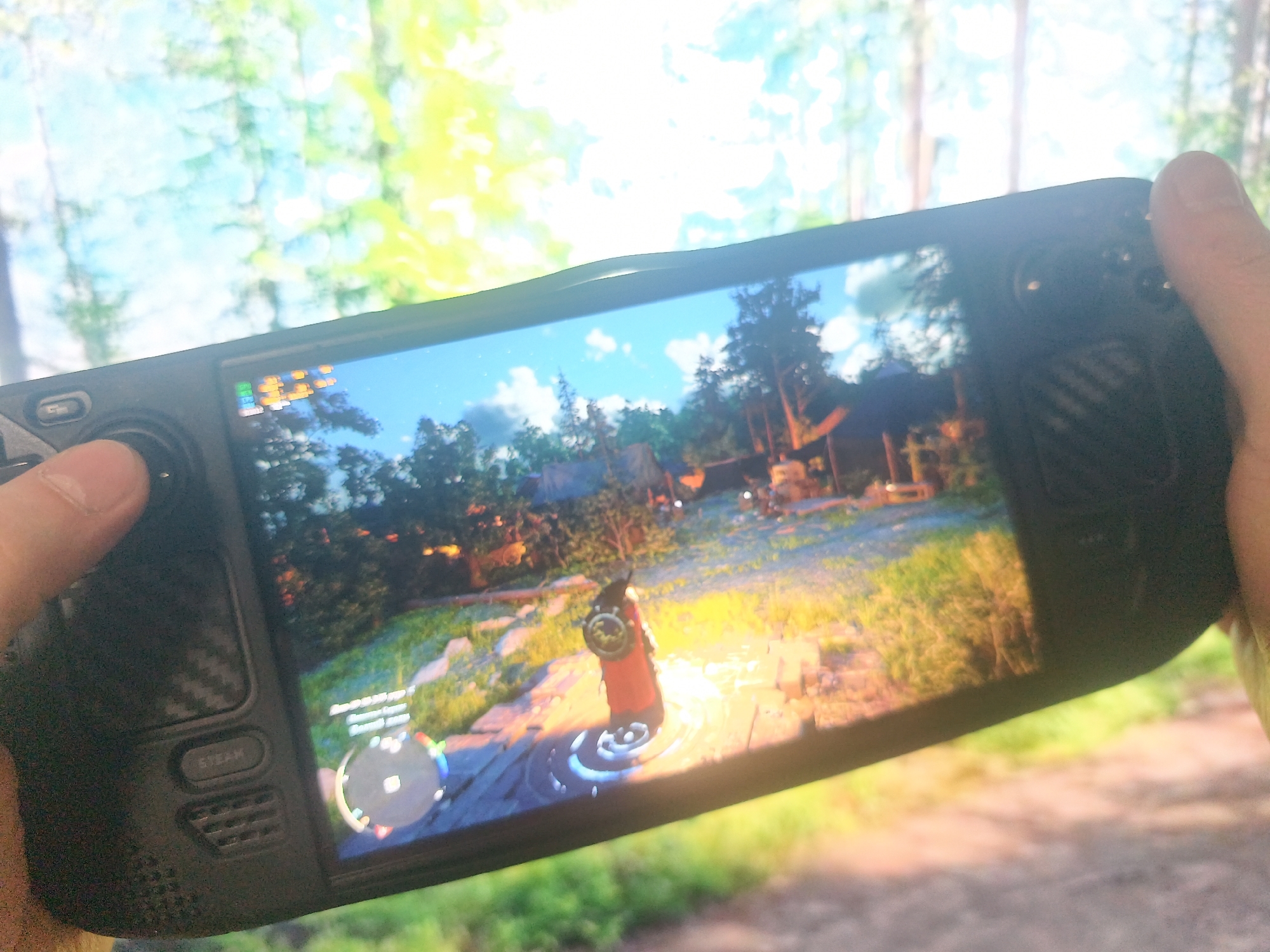 A handheld gaming device, likely a Steam Deck, is being held up in front of a blurred outdoor scene. The screen displays a video game with a third-person perspective, showing a character in a red robe standing on a circular platform in a grassy environment with trees and buildings in the background. The device is black and has visible buttons and vents. The background appears to be a natural setting with trees and foliage, but the details are indistinct due to the blur. The precise location within Chaikovsky, Russia, cannot be determined from the image.