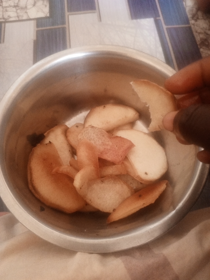 The image shows a metallic bowl, viewed from above, containing numerous fried potato pieces or wedges. The potato pieces vary in color from light golden-brown to paler shades, with some exhibiting reddish remnants of potato skin and small dark specks. A dark-skinned human hand is positioned on the right side of the bowl, holding a single, broken piece of fried potato between its thumb and index finger. The bowl rests on a light-colored, subtly striped fabric surface. In the background, an upright tiled surface with a geometric pattern of light and dark blue-gray rectangular and square sections is visible. The scene is consistent with an indoor, casual eating environment within the city of Kuta, Nigeria.