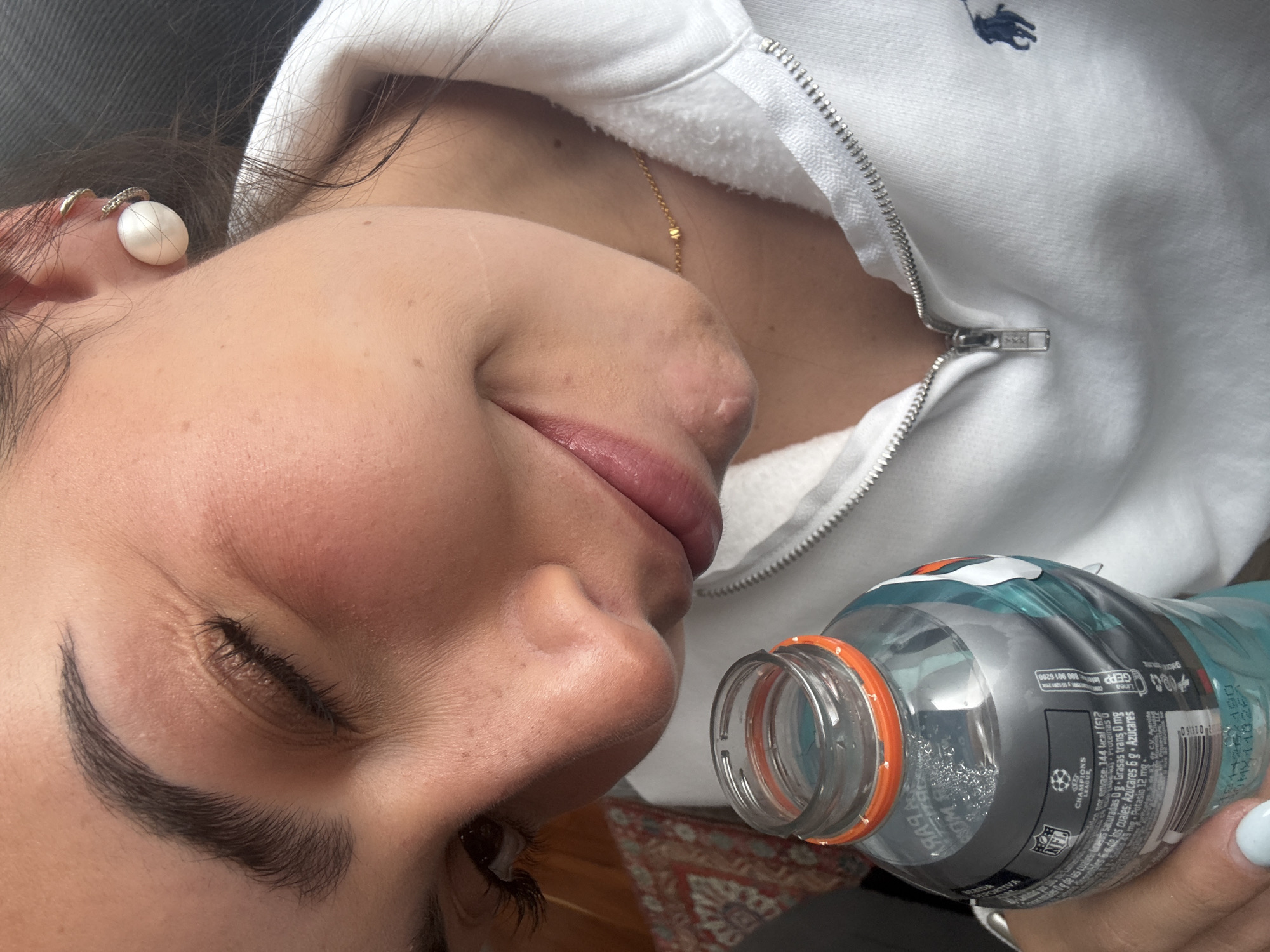 A young woman is shown from the side, appearing to be in a reclined position. She is wearing a white zip-up hoodie with a logo on the upper right side, as well as a gold necklace and pearl earring. She holds a bottle of Gatorade, obscuring the lower part of her face. The Gatorade bottle is slightly open. A portion of a rug is visible in the lower part of the image. The location in San Bernardino Tlaxcalancingo, Mexico cannot be determined from the image alone.